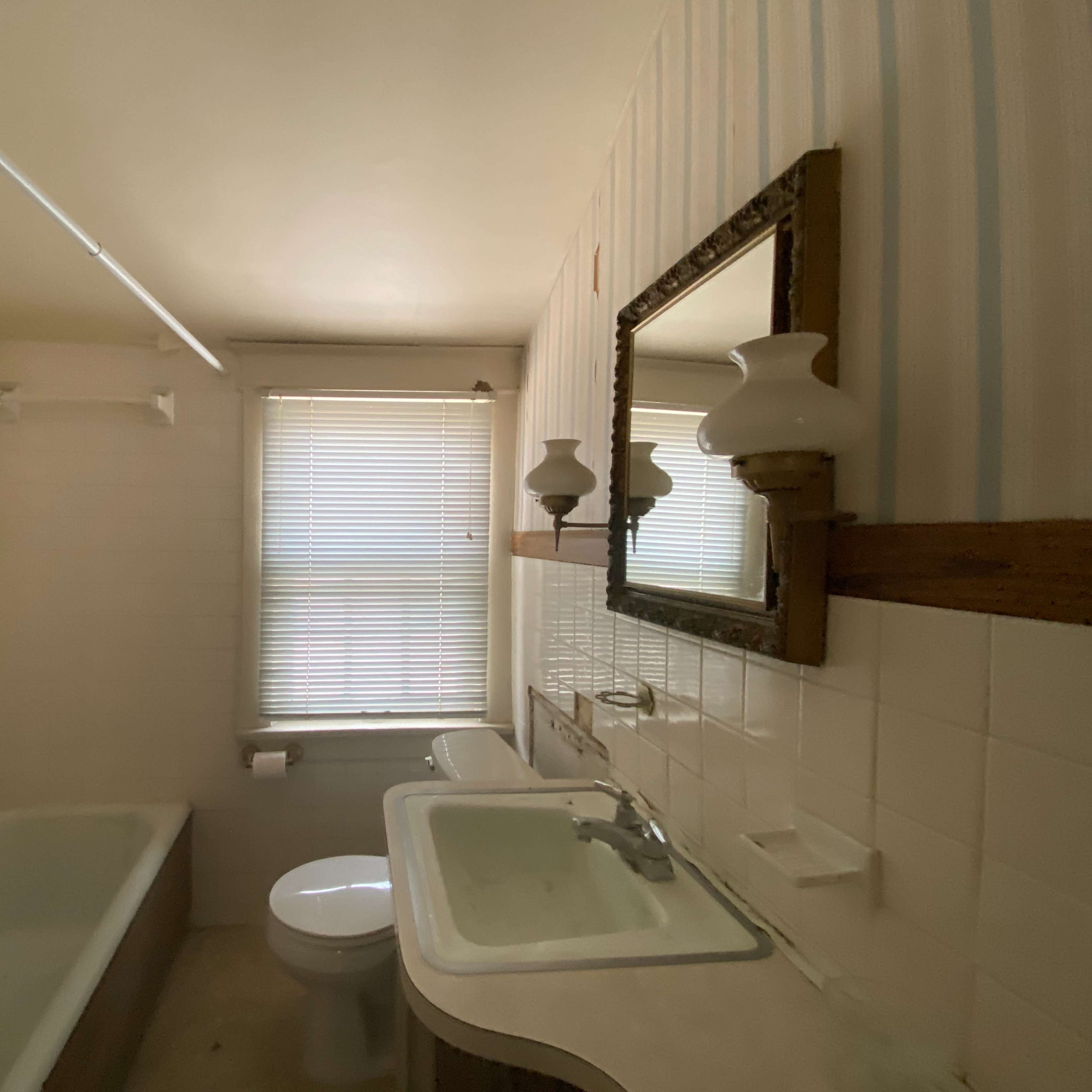 A bathroom with a bathtub, toilet, sink, and a mirror, featuring wallpaper and window blinds.