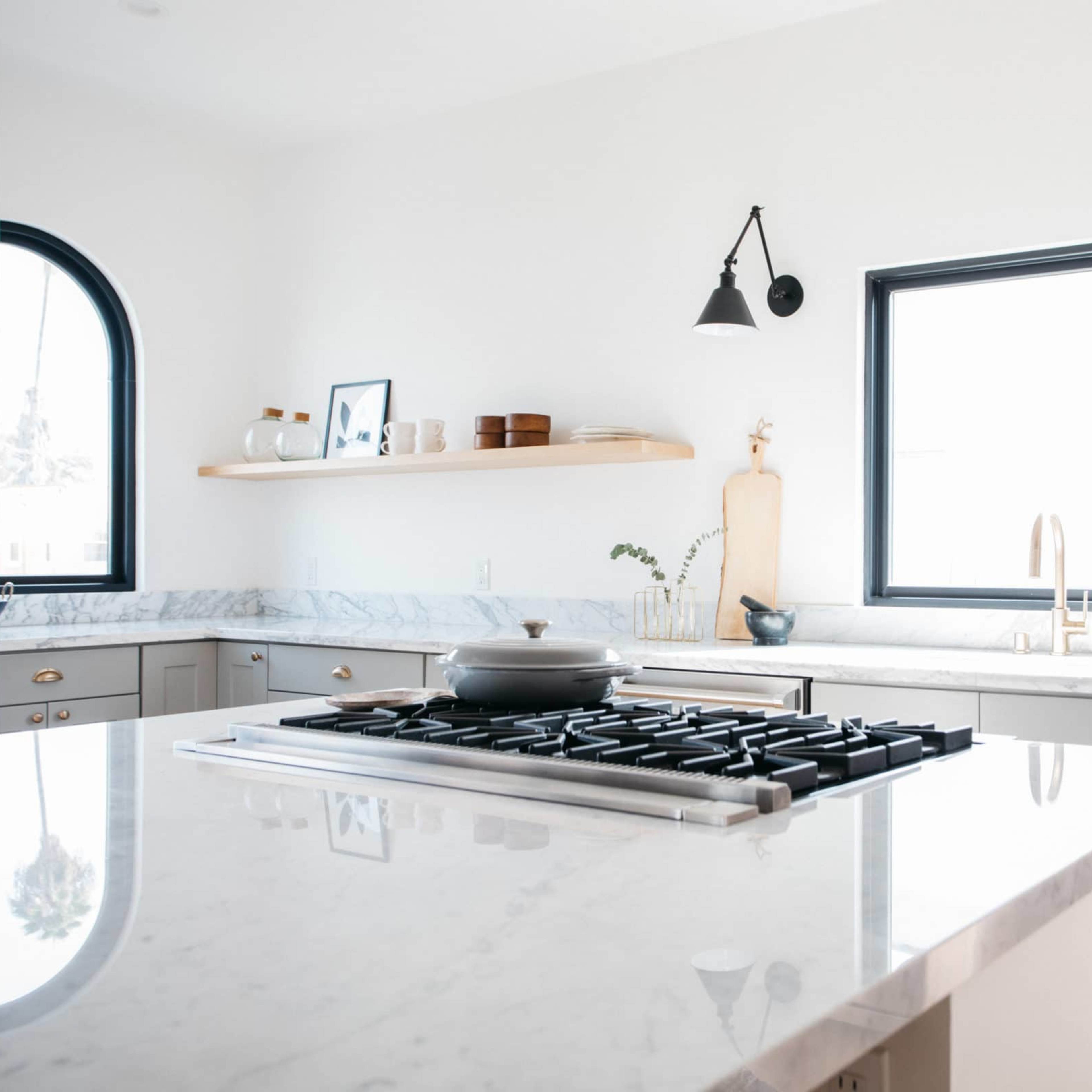 A modern kitchen features a central island with a gas stove, marble countertops, and minimalist decor against white walls and black-framed windows.