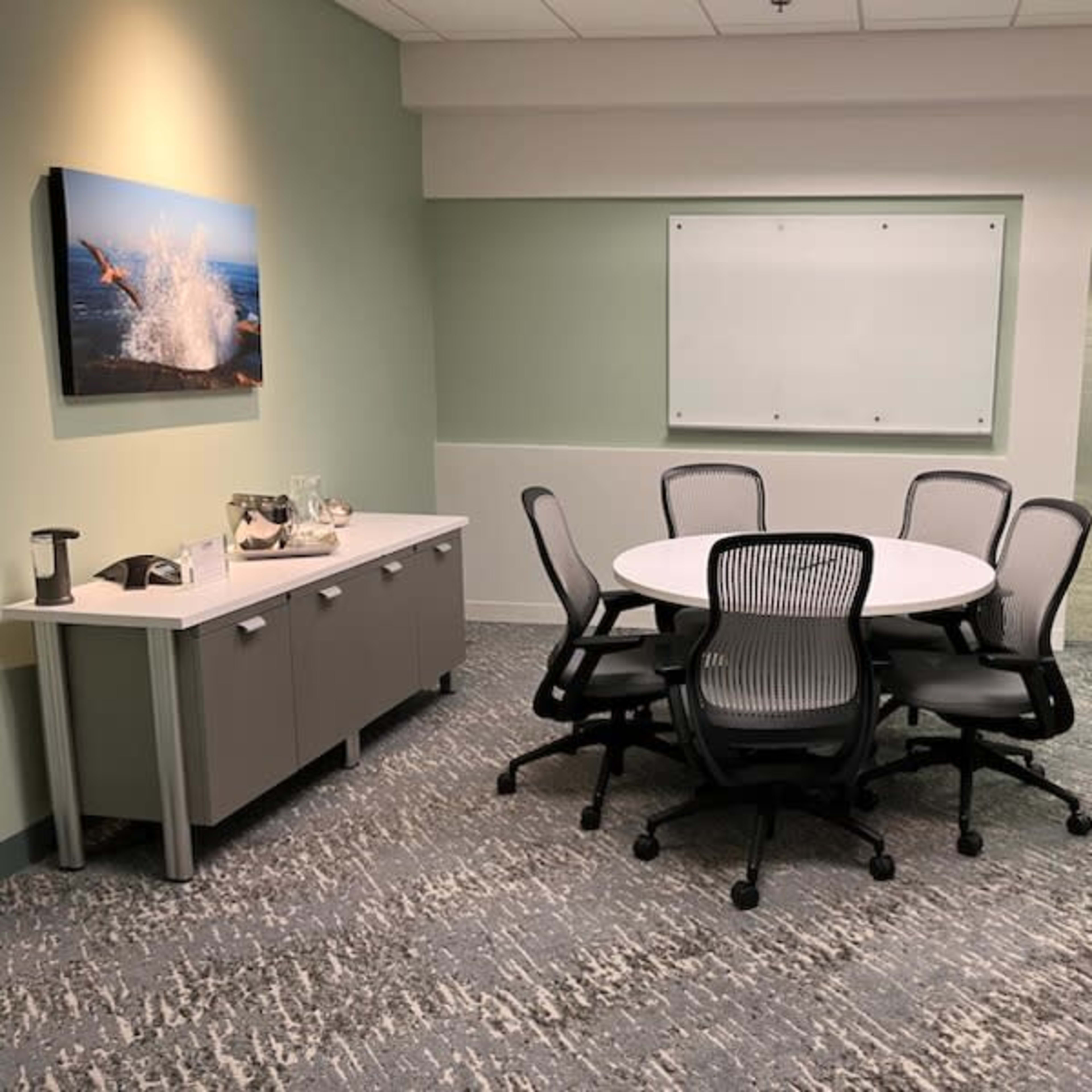 A small meeting room with a round table surrounded by five chairs, a side cabinet with a beverage setup, and a whiteboard on the wall.