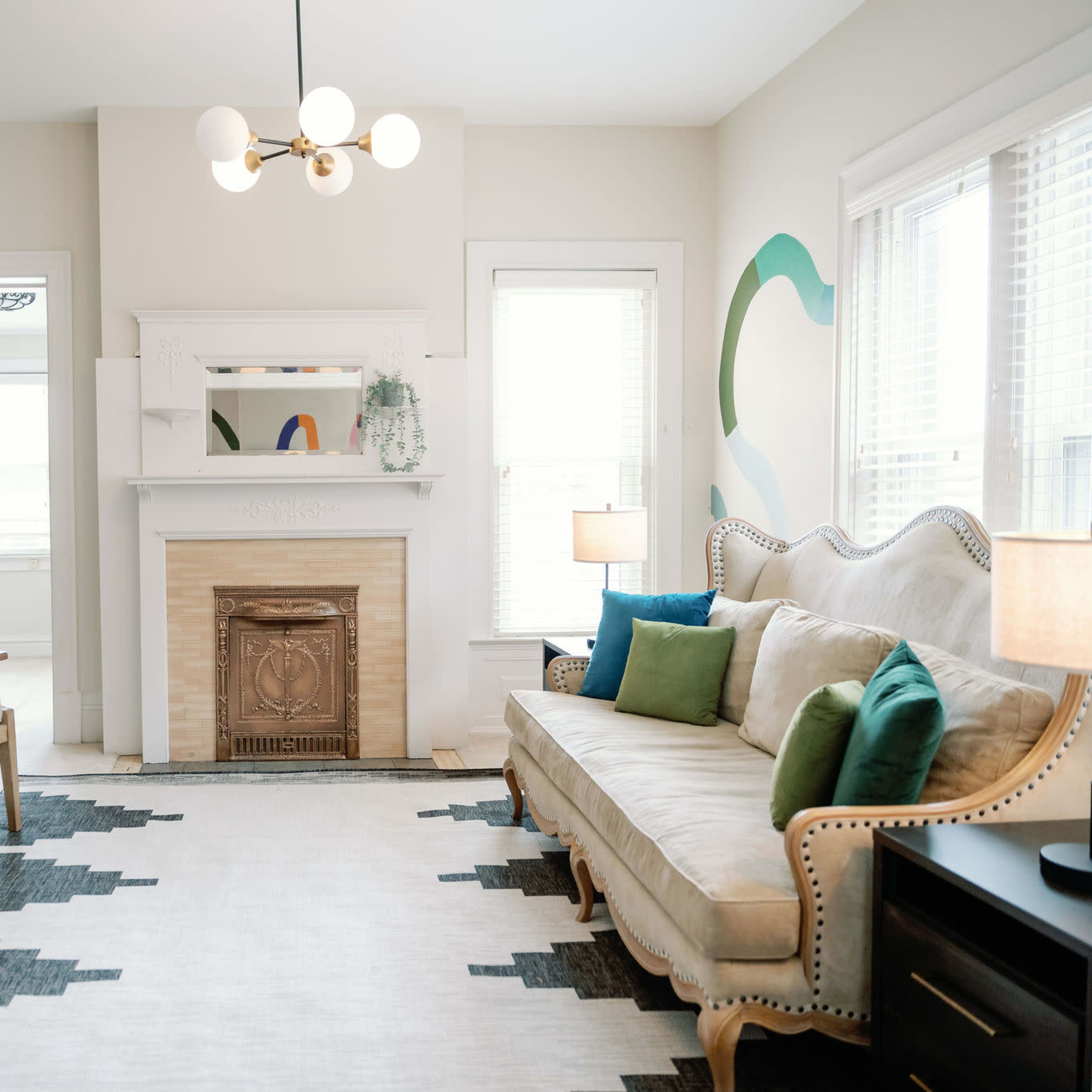A cozy living room with a light-colored sofa, two wooden armchairs, a fireplace, and decorative pillows arranged around a patterned rug.