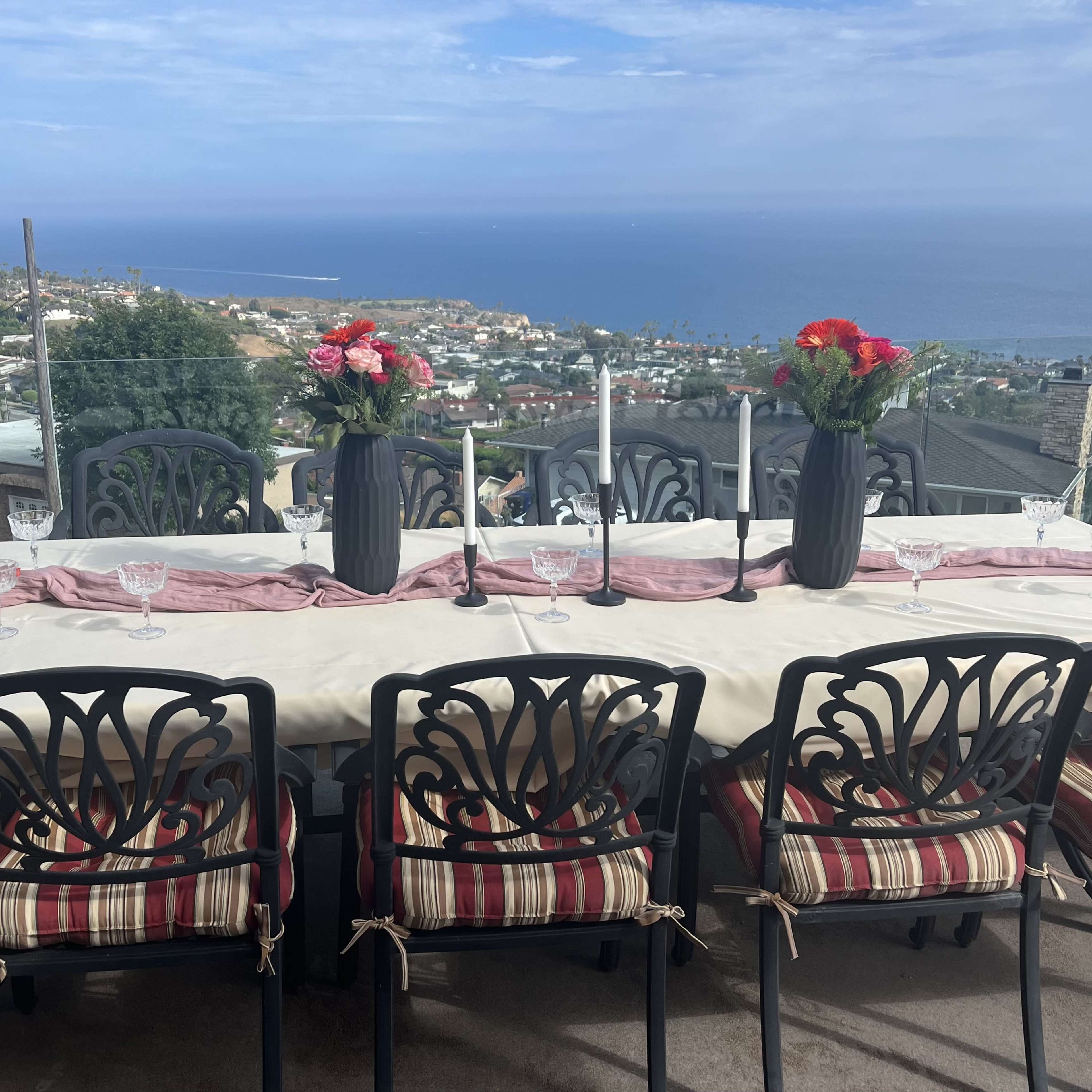 A long dining table is set with place settings and floral centerpieces overlooking a coastal view.