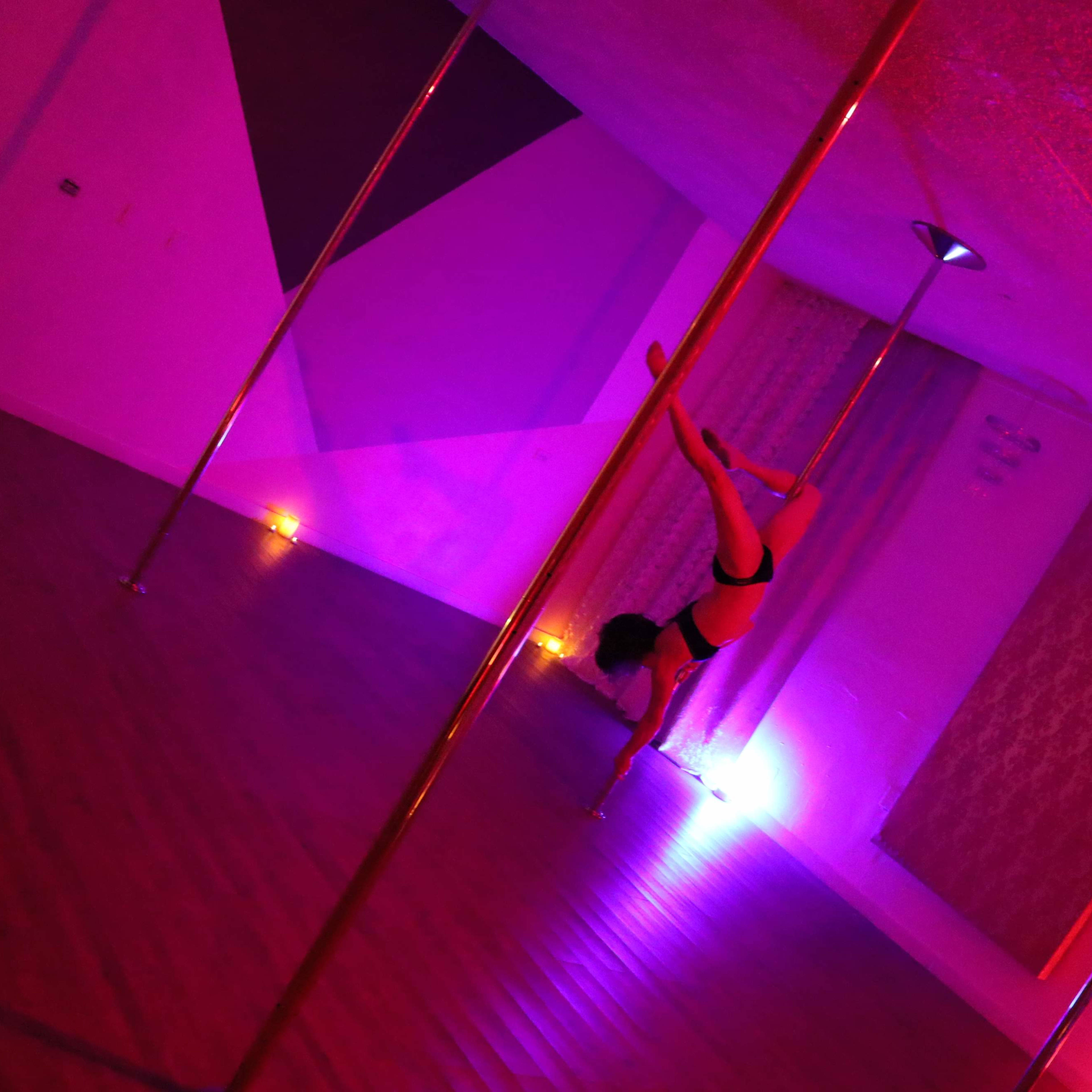 A dancer performs a handstand on a pole in a dimly lit room with purple and pink lighting.