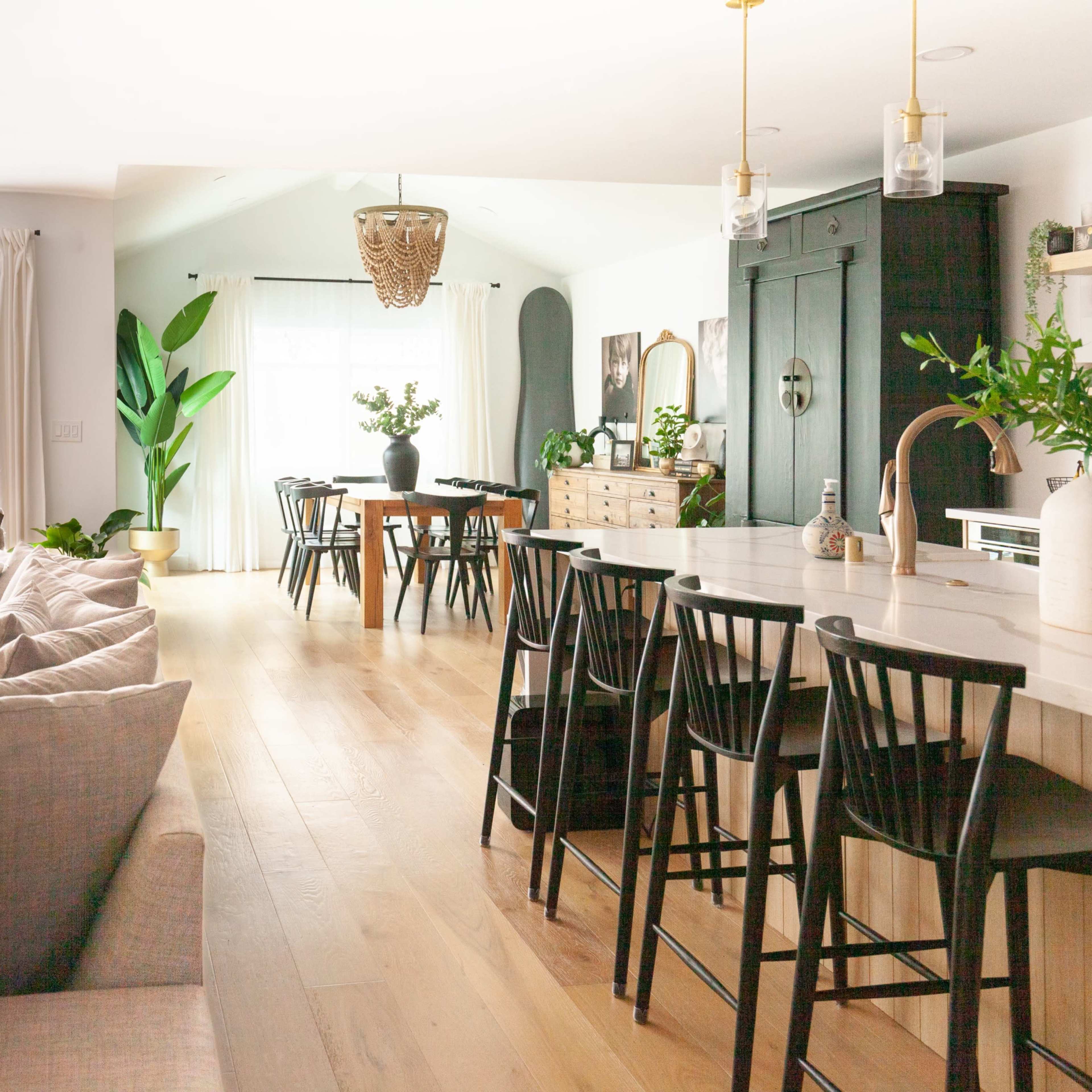 A bright and open living space features a kitchen island with bar stools, a dining table, and a lounge area with a sectional sofa, leading to a pool area visible through sliding glass doors.