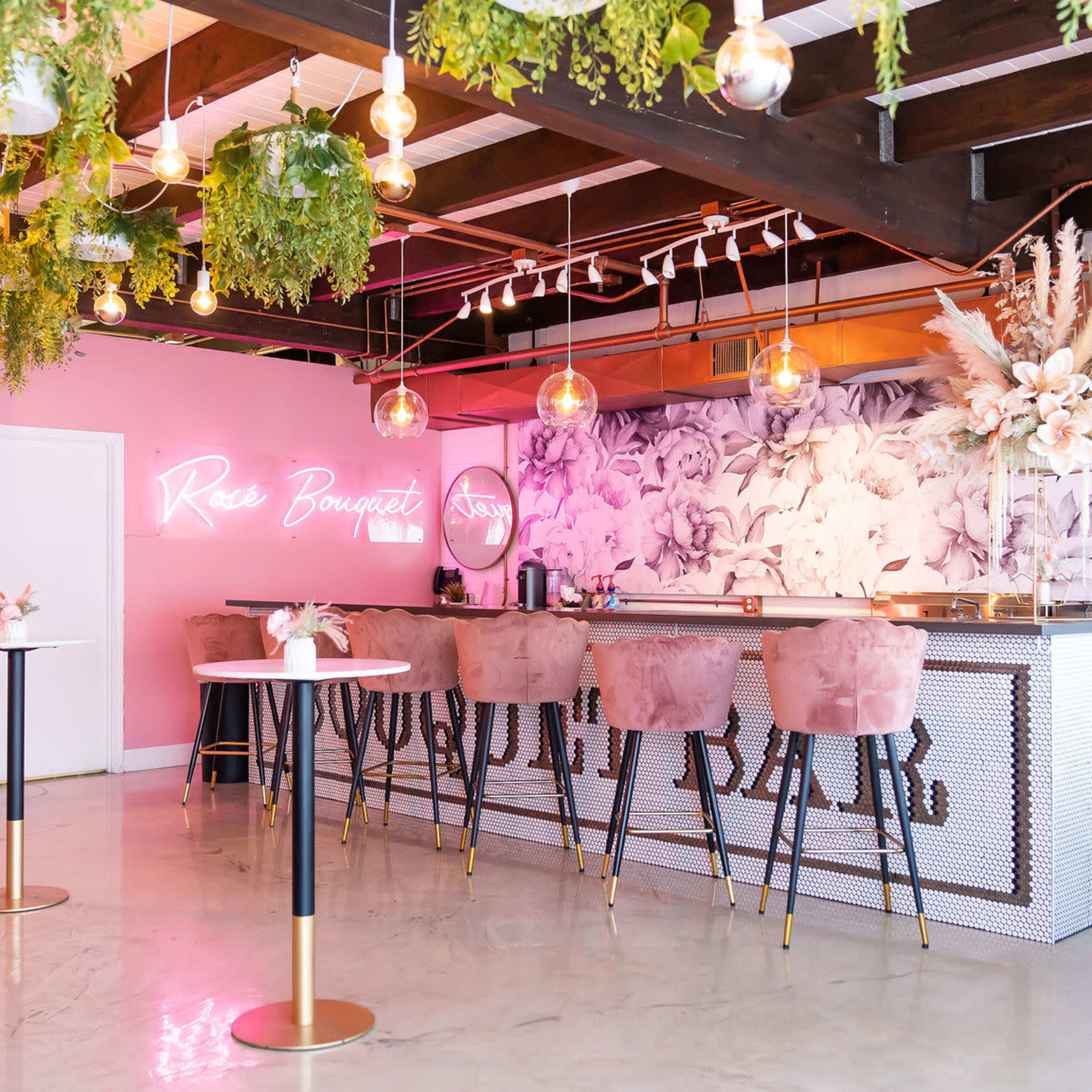 A brightly adorned bar area with pink seating, greenery hanging from the ceiling, and a floral mural on the wall.