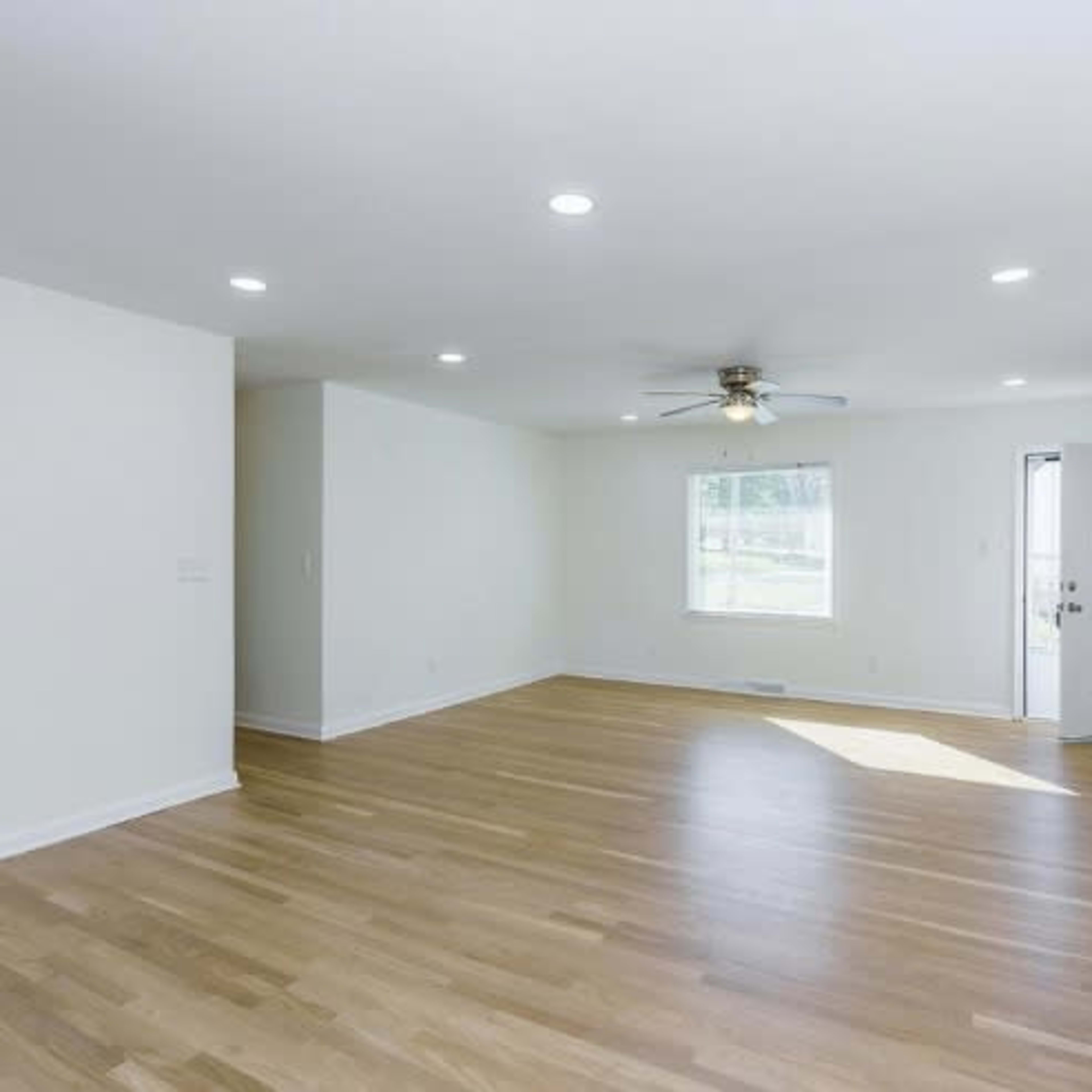 A spacious, empty living room with light-colored walls and wooden flooring, featuring a ceiling fan and a window next to a door.