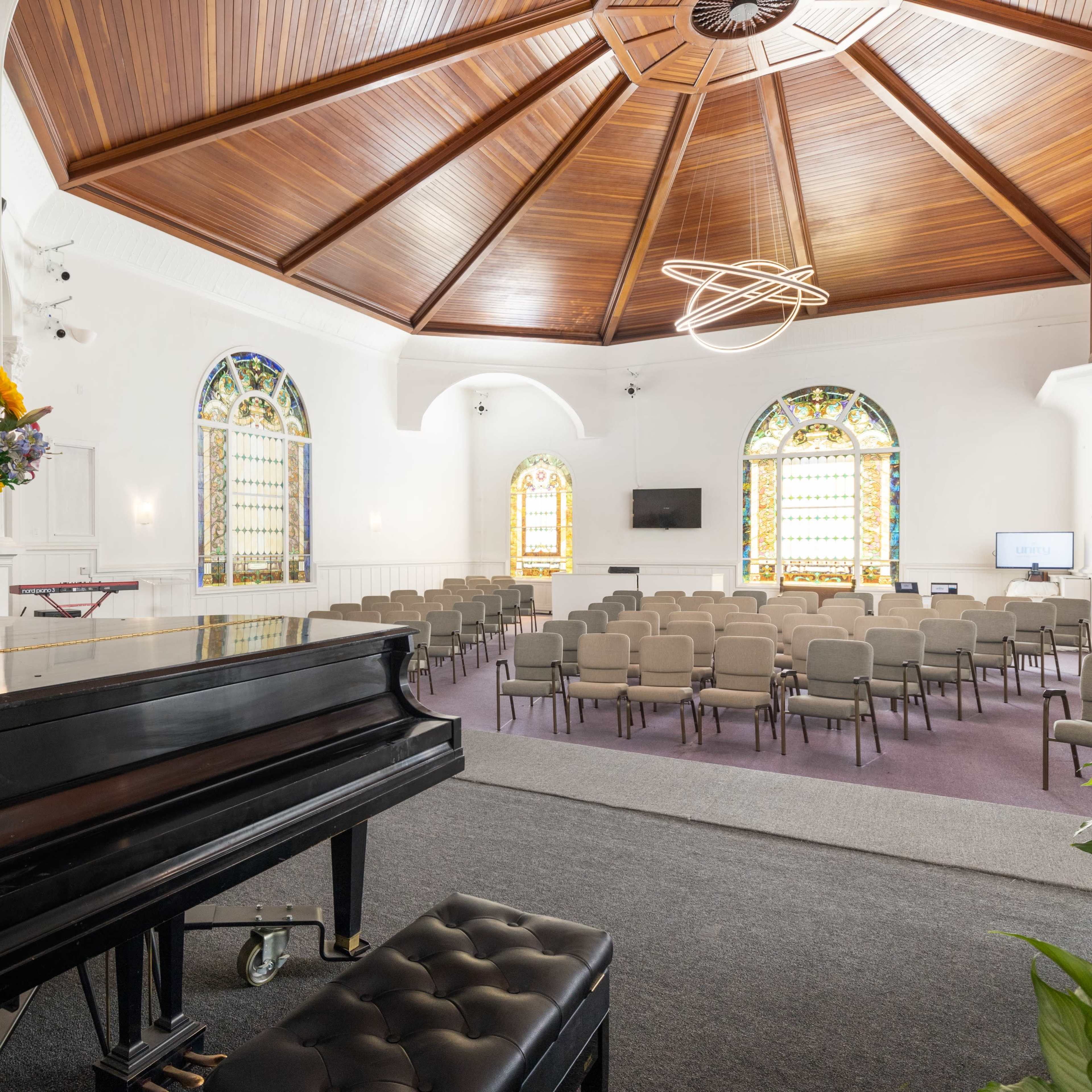 A spacious church interior featuring a grand piano, rows of chairs, and stained glass windows under a wooden vaulted ceiling.