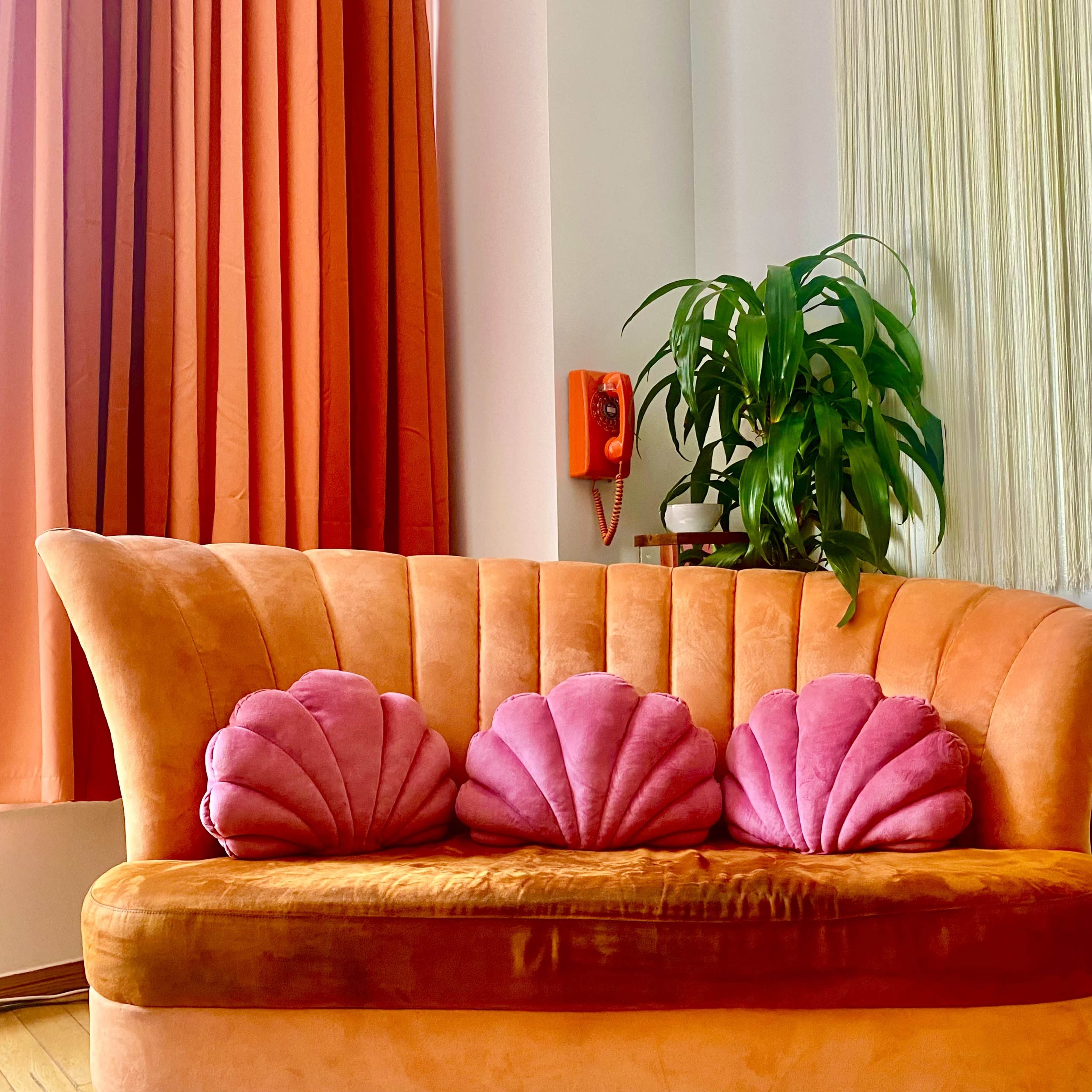 A curved orange sofa with three pink shell-shaped cushions sits against a backdrop of colorful curtains and a houseplant.