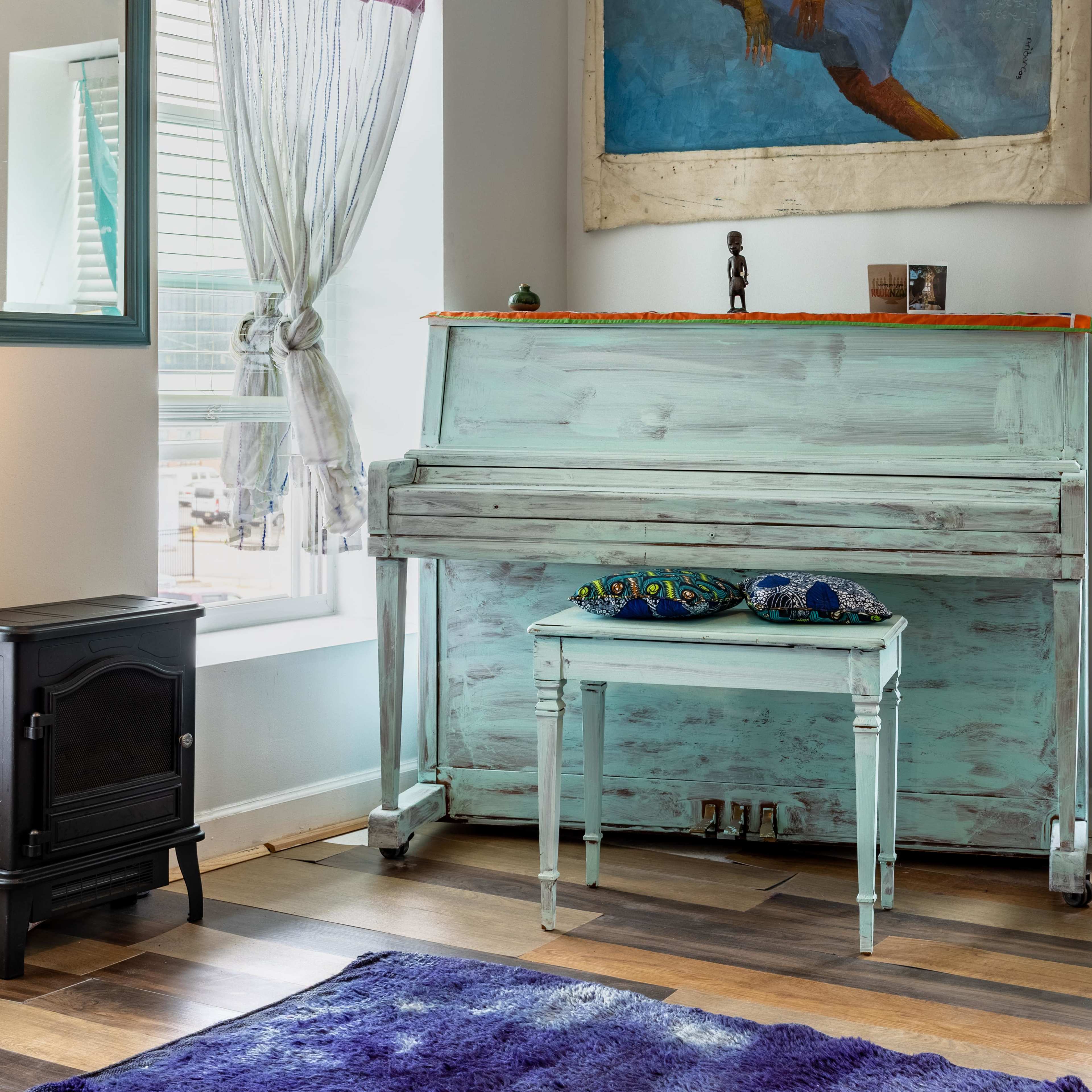A light blue piano stands against a wall, accompanied by a small table and a woven basket, with natural light streaming through the windows.