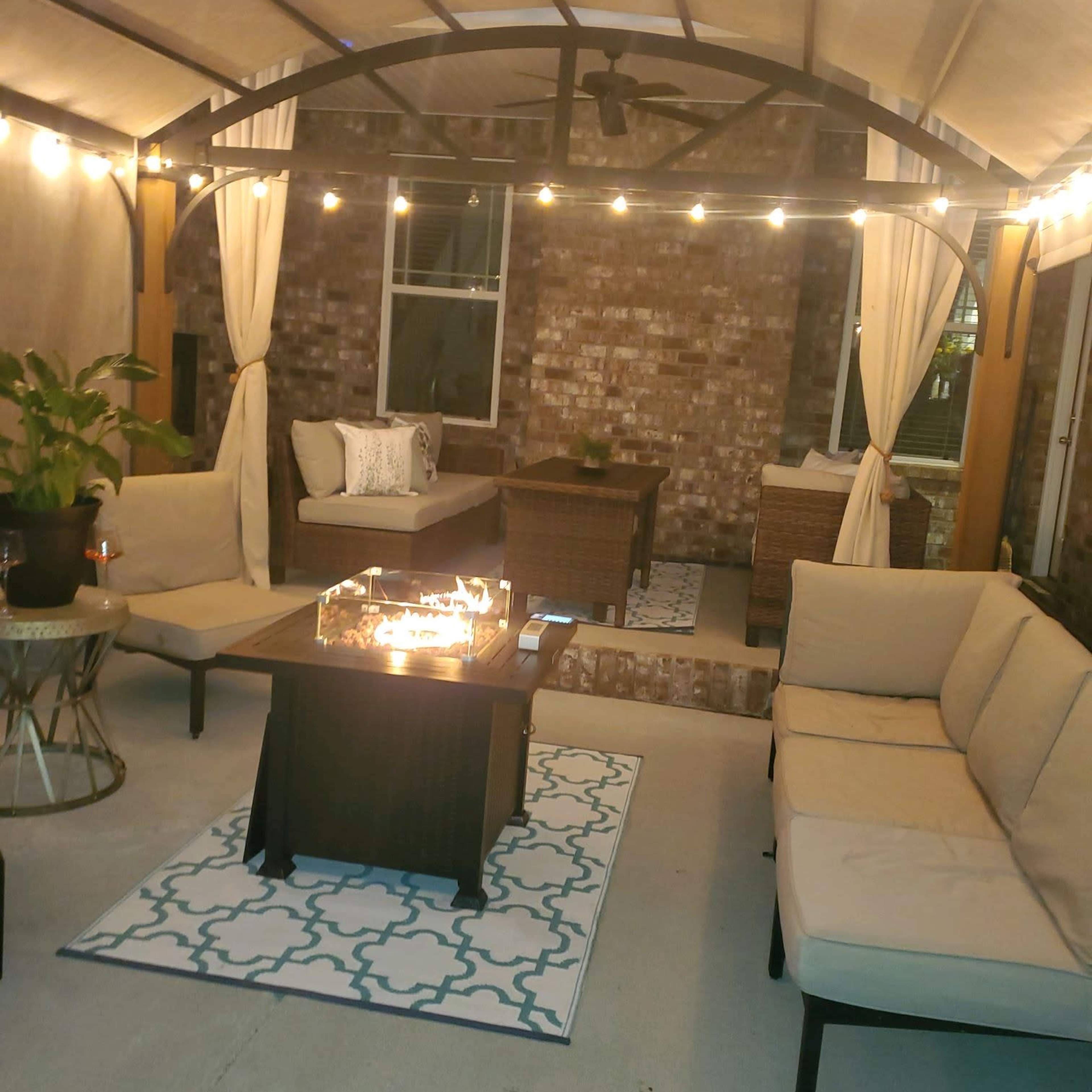 A cozy outdoor seating area under a canopy with string lights, featuring a fire pit table surrounded by cushioned chairs and a sofa.