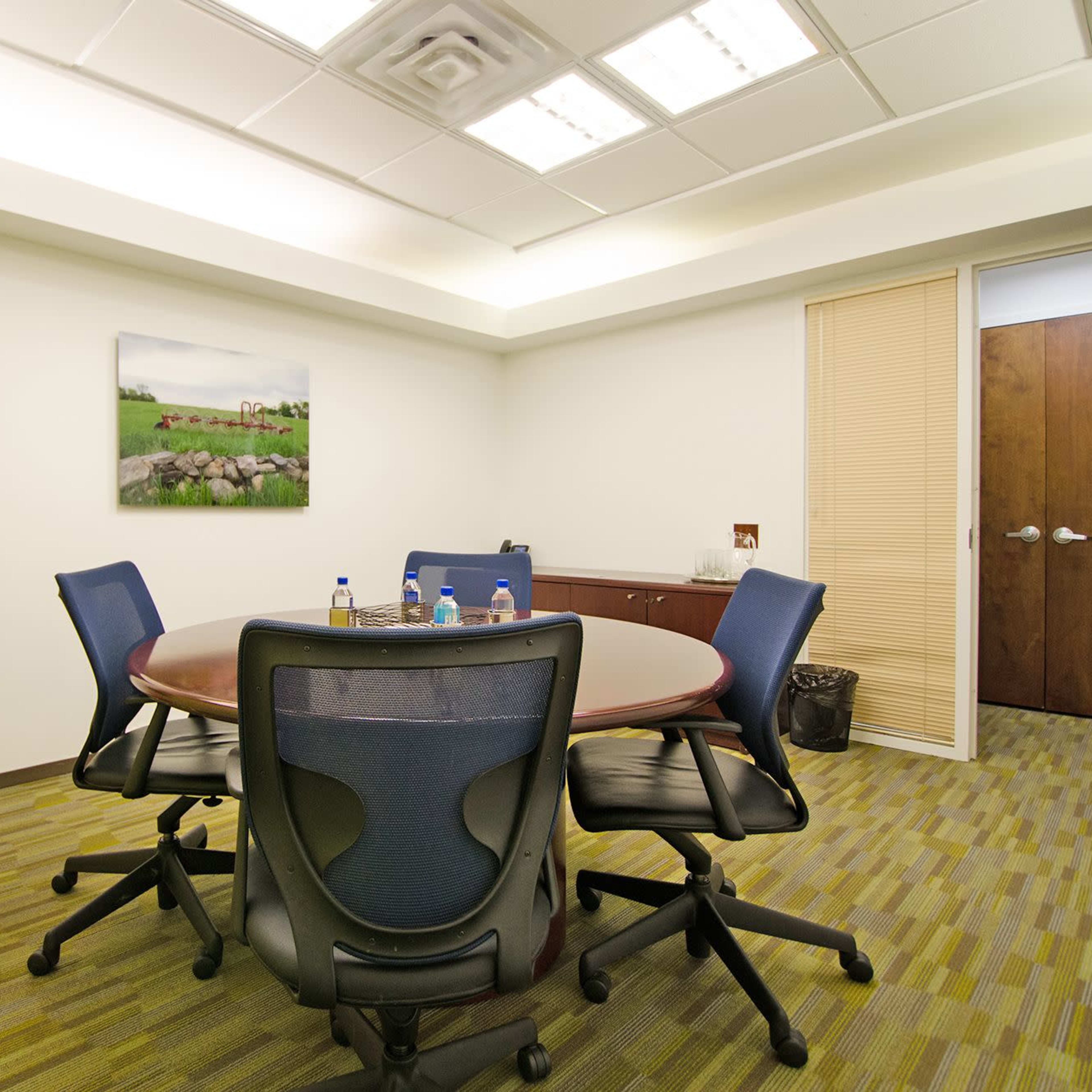 A conference room features a round table with four chairs, a plant, and a piece of artwork on the wall.