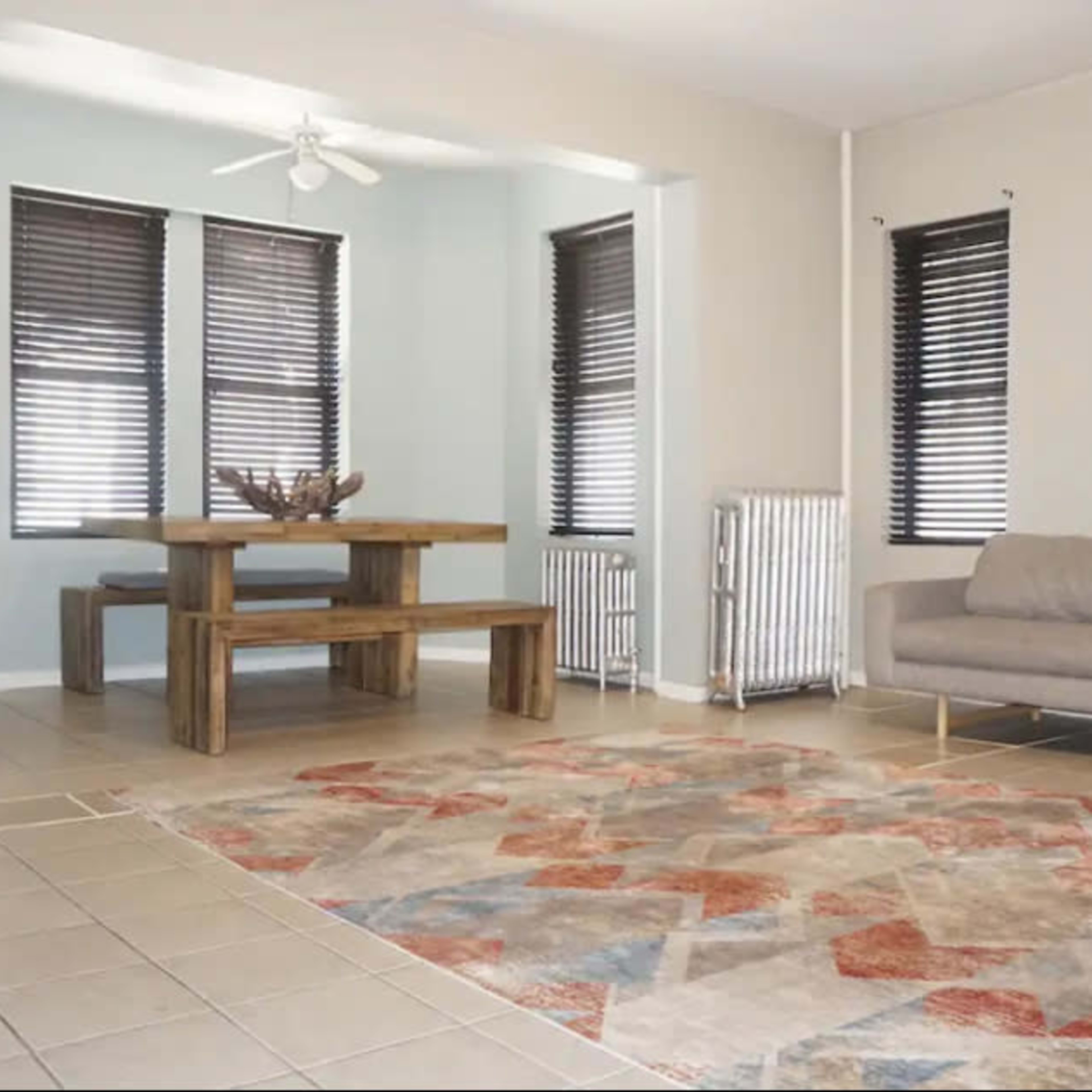 A spacious room featuring a wooden dining table, a gray sofa, a patterned area rug, and large windows with dark blinds.