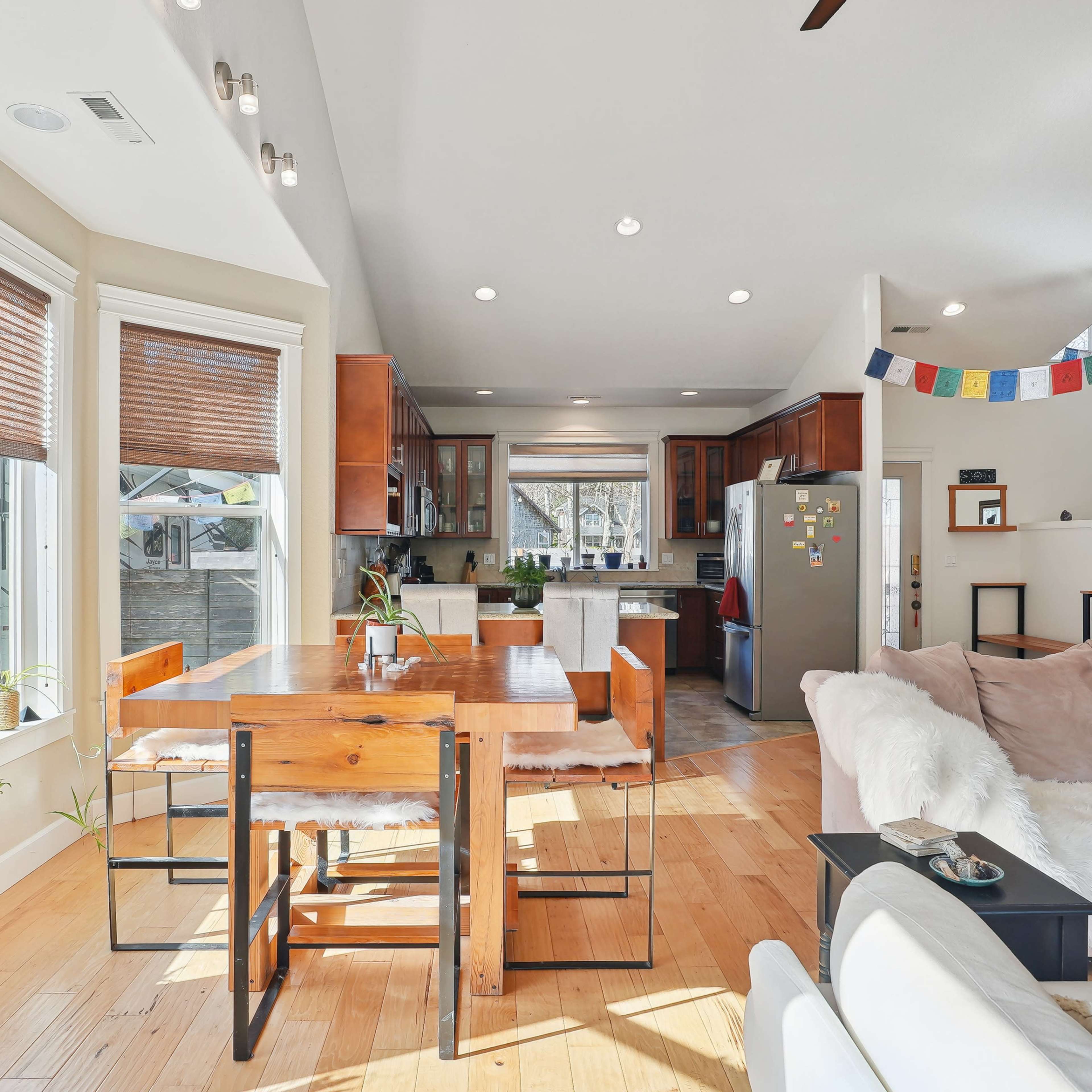 A modern living space features a dining area with a wooden table and chairs, surrounded by large windows, open to a kitchen that includes a refrigerator and wood cabinets.