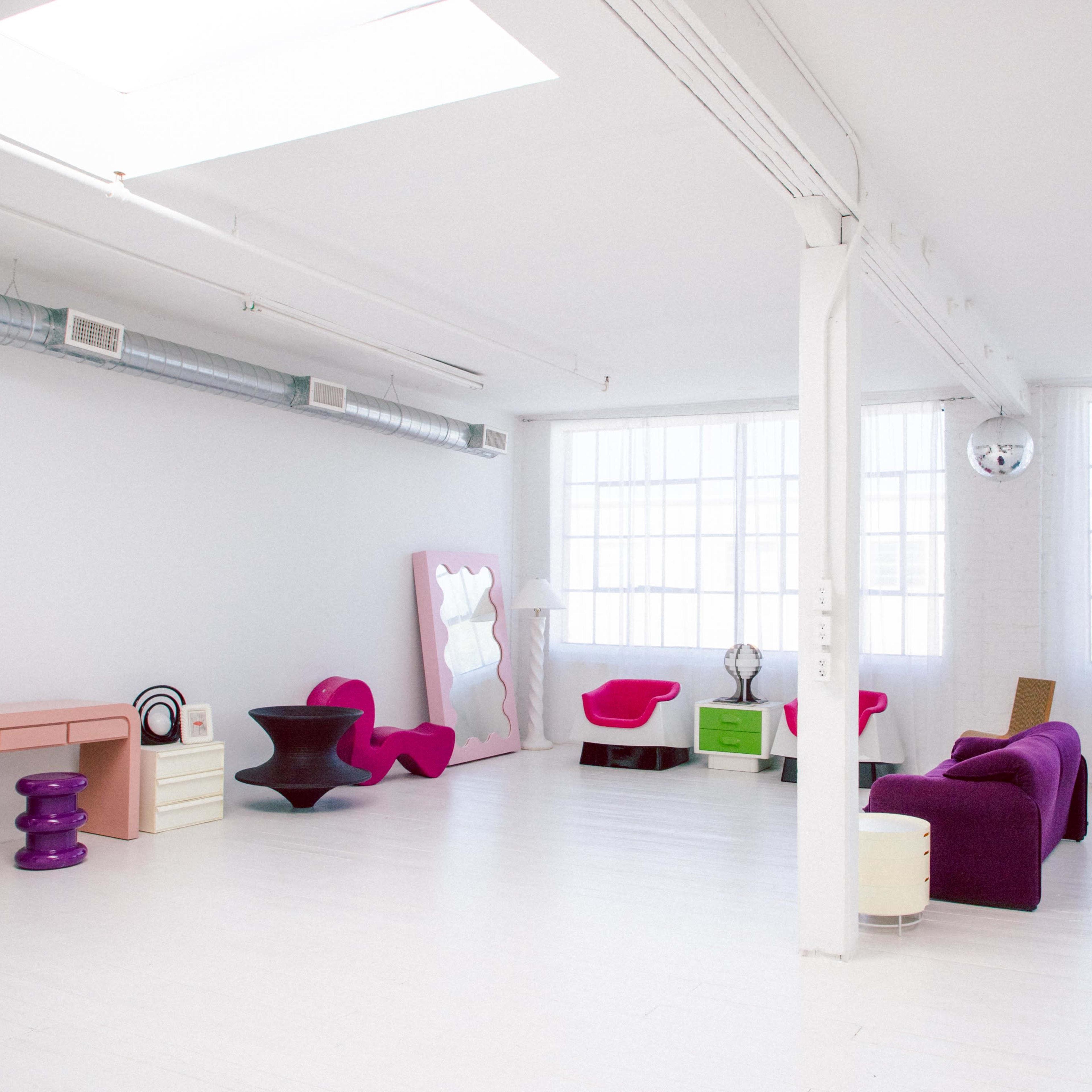 A spacious, well-lit room featuring colorful modern furniture, including a pink sofa, teal seating, and a unique purple table, against a backdrop of white walls and large windows.