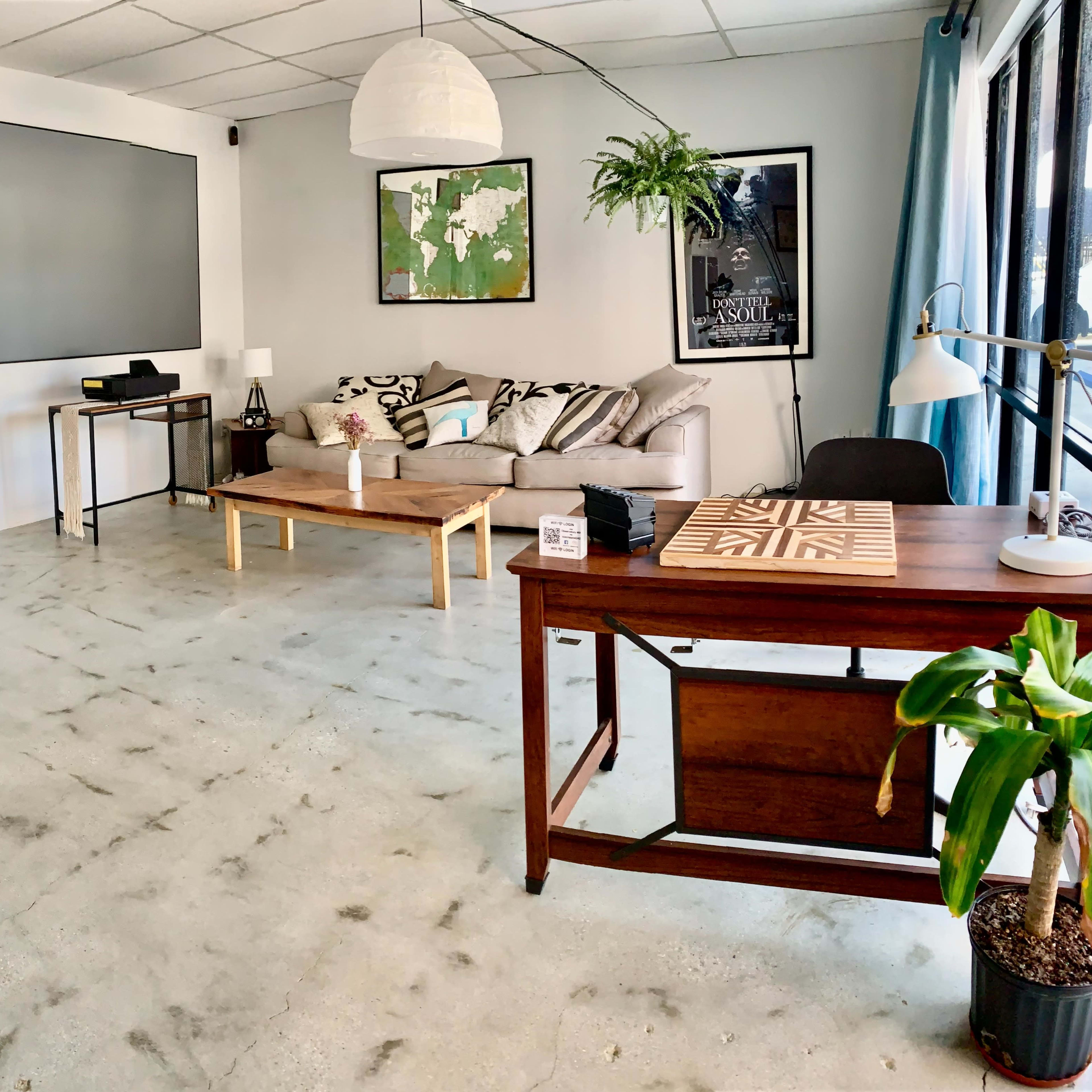 A modern office space features a large sofa, a coffee table, a desk, and a wall-mounted screen.