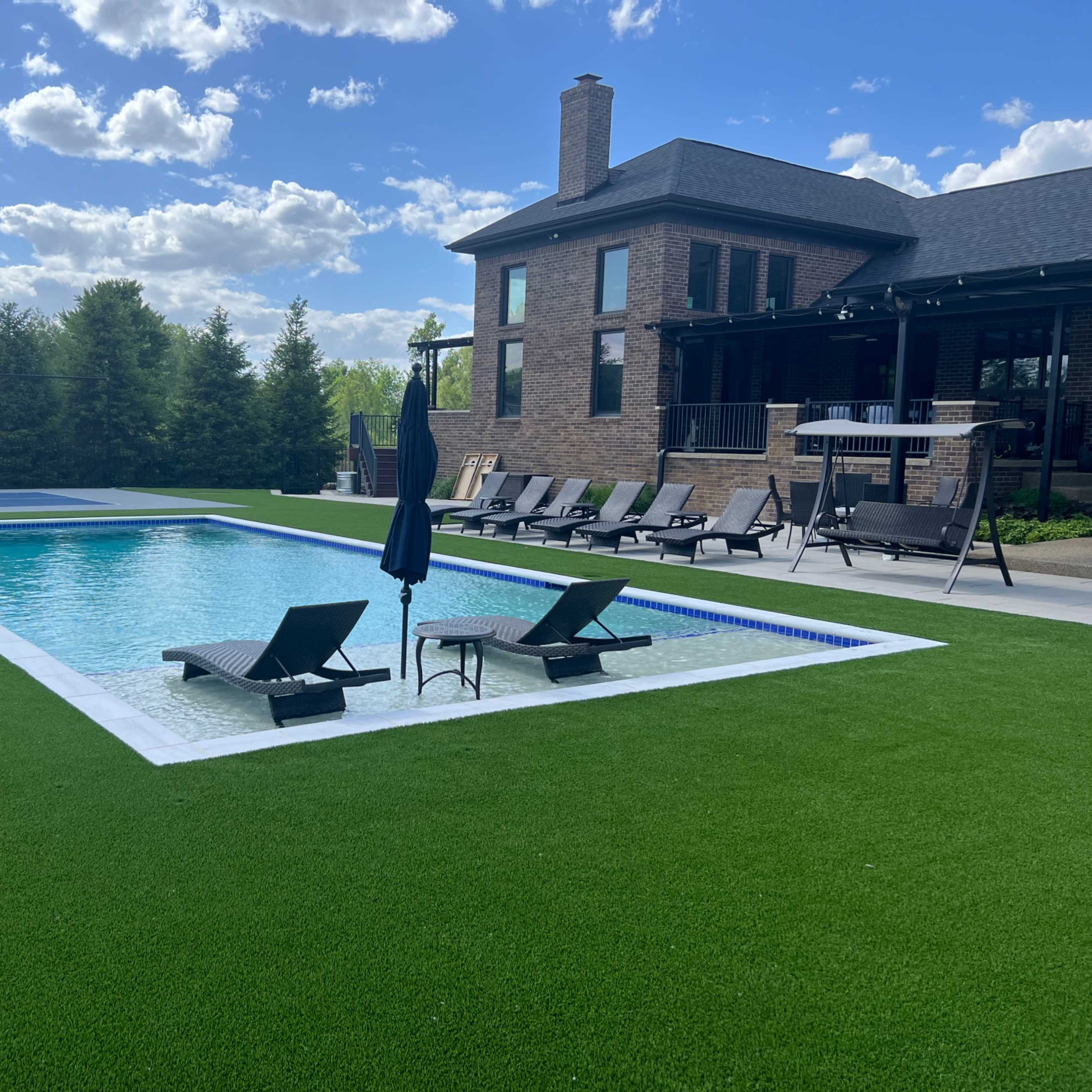 A backyard with a paved swimming pool surrounded by lawn chairs, a basketball court in the background, and a two-story brick house.