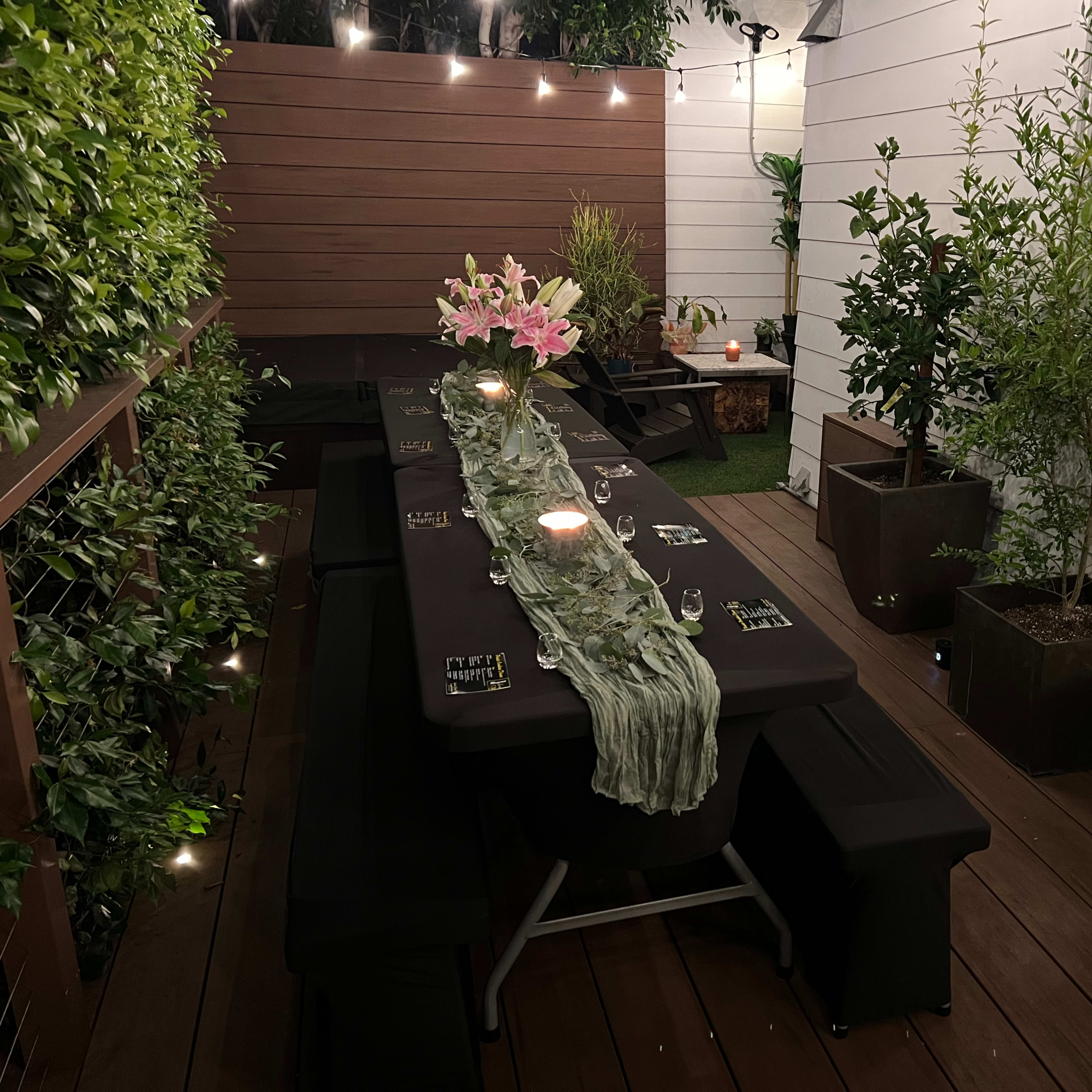 A long table is set for a dinner in a well-lit outdoor patio, surrounded by greenery and decorative potted plants.