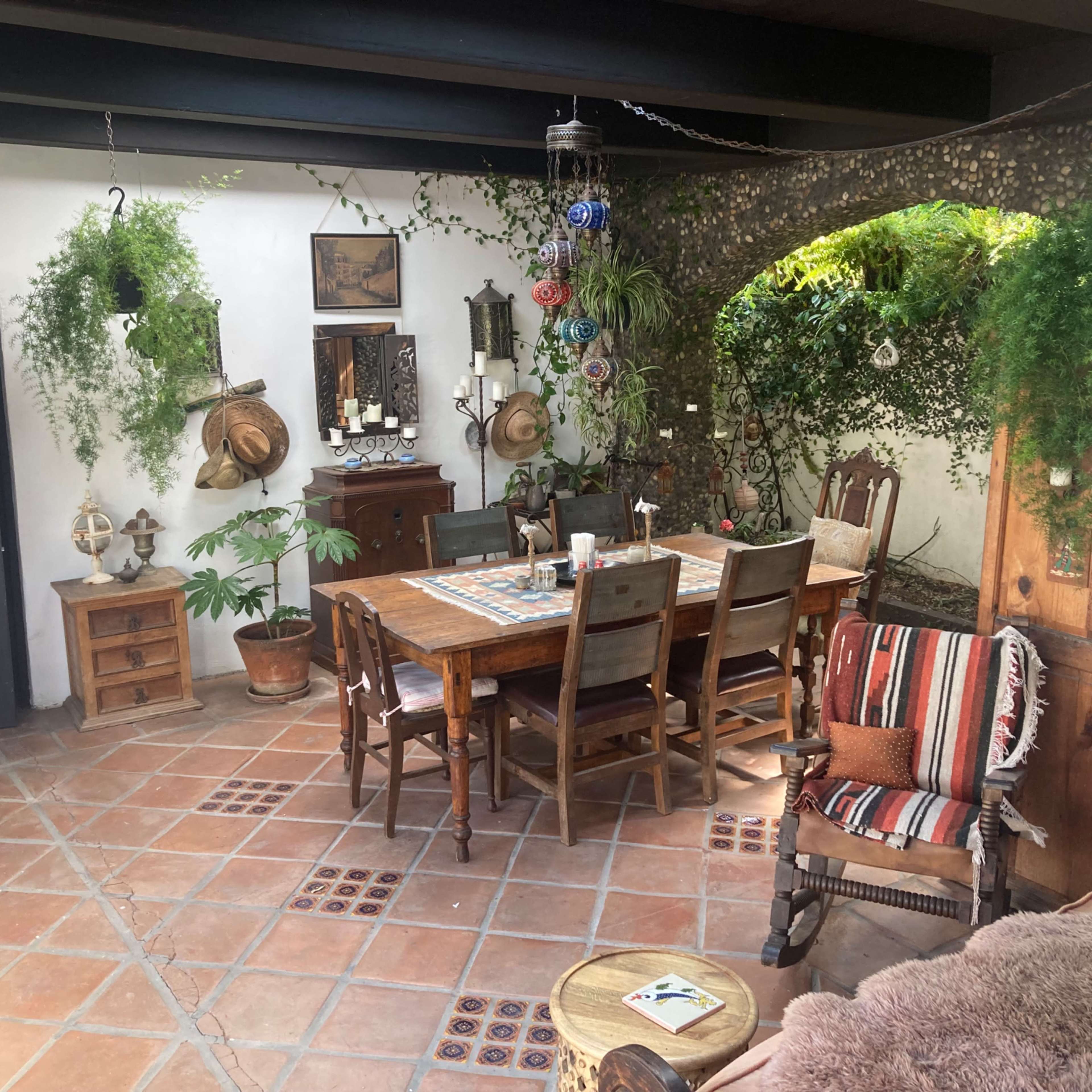 A cozy dining area with a wooden table surrounded by chairs, decorative plants hanging from the ceiling, and various pieces of rustic furniture.