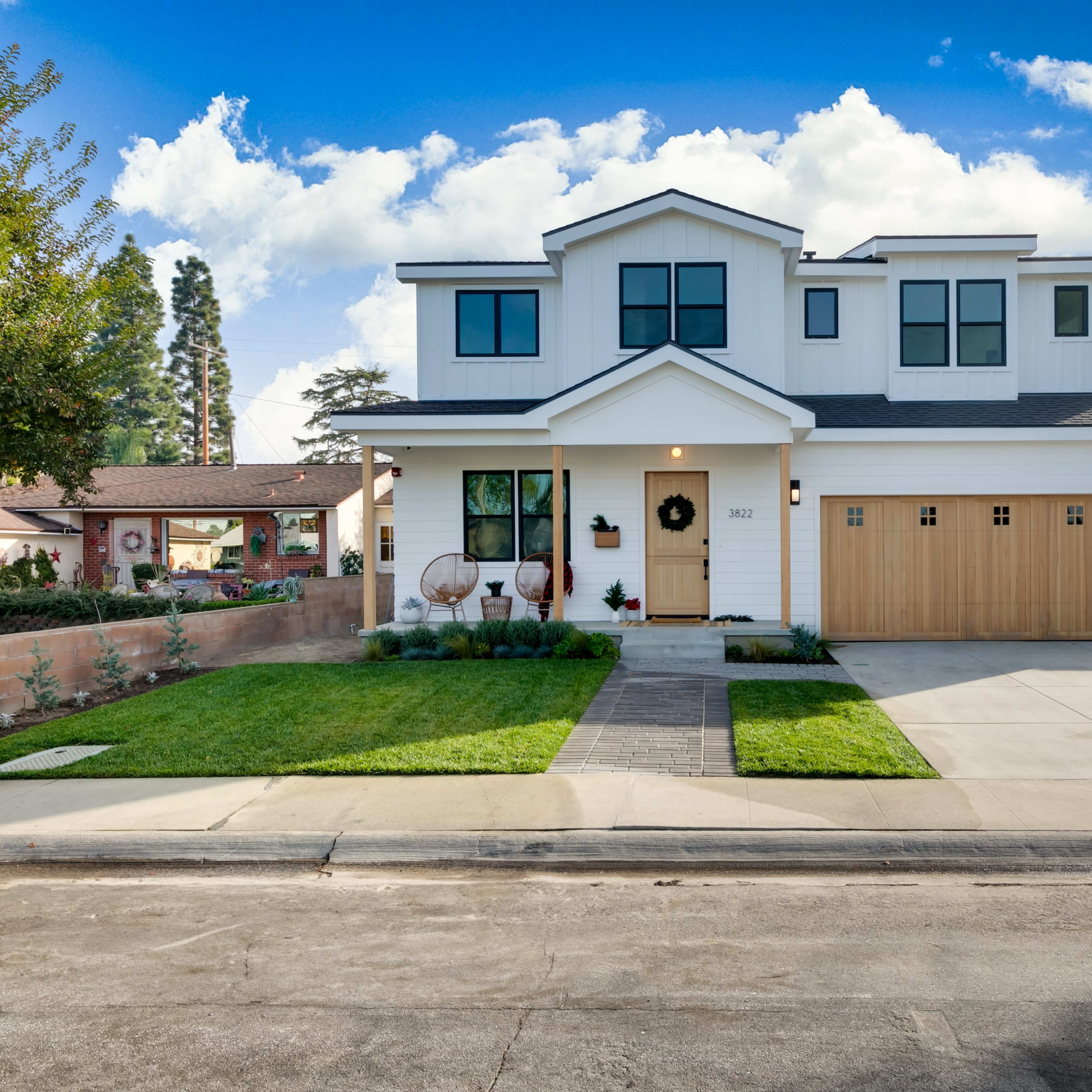 A modern two-story house with a white exterior and a wooden garage door is situated on a residential street, featuring a landscaped front yard and a paved driveway.