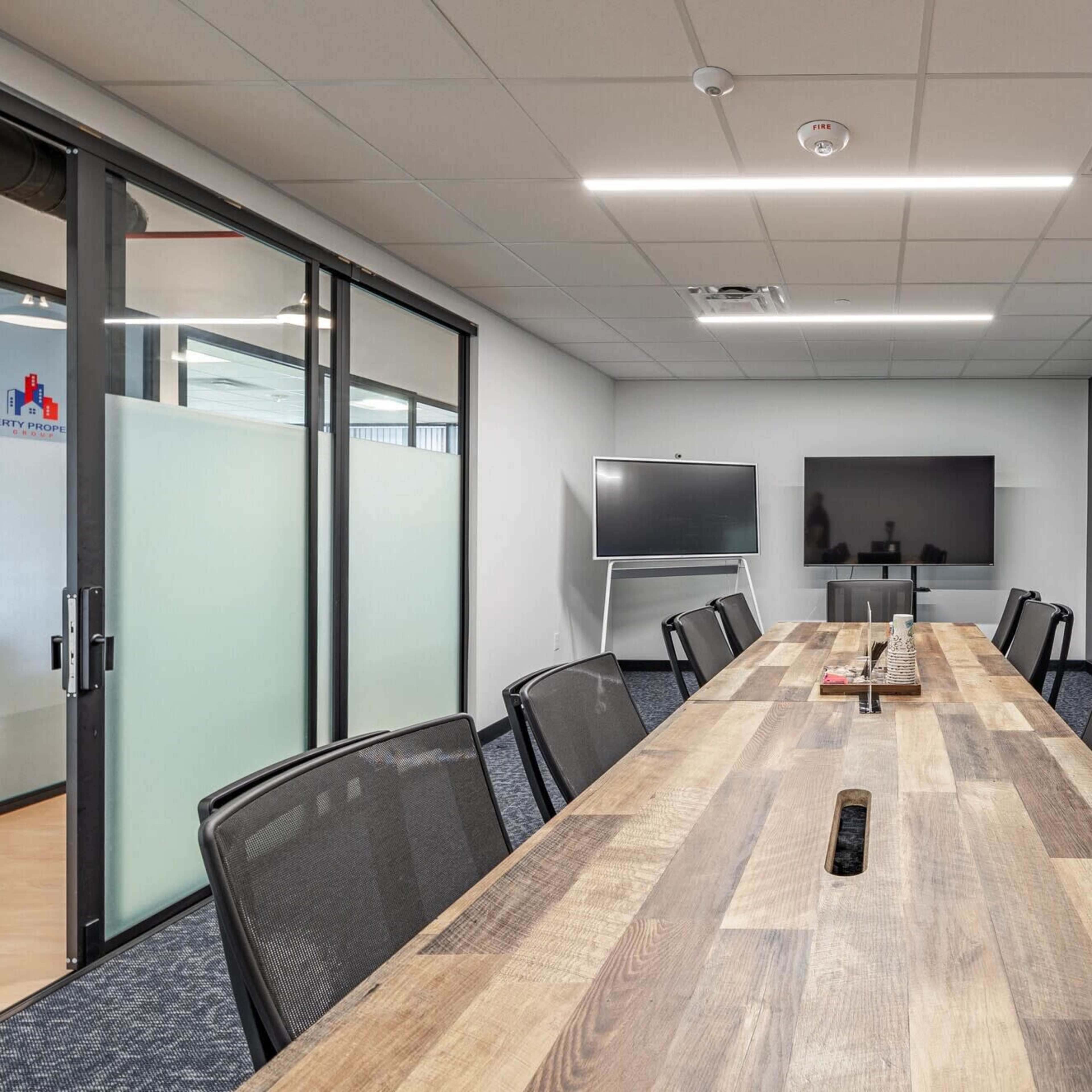 A modern conference room with a long wooden table, black mesh chairs, two large screens, and whiteboards on the walls.