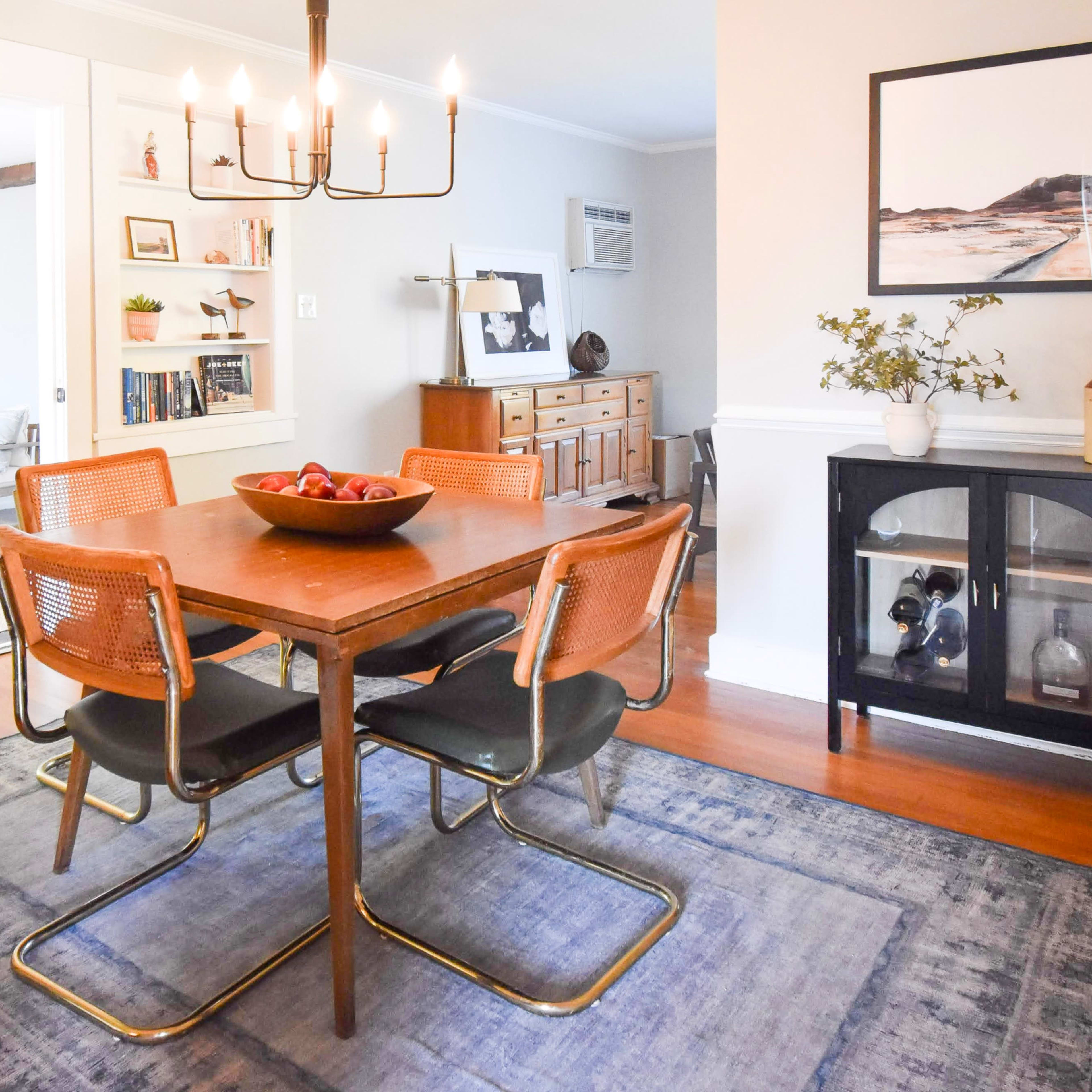 A dining room features a wooden table surrounded by four chairs, with a chandelier above and a bowl of fruit as a centerpiece.