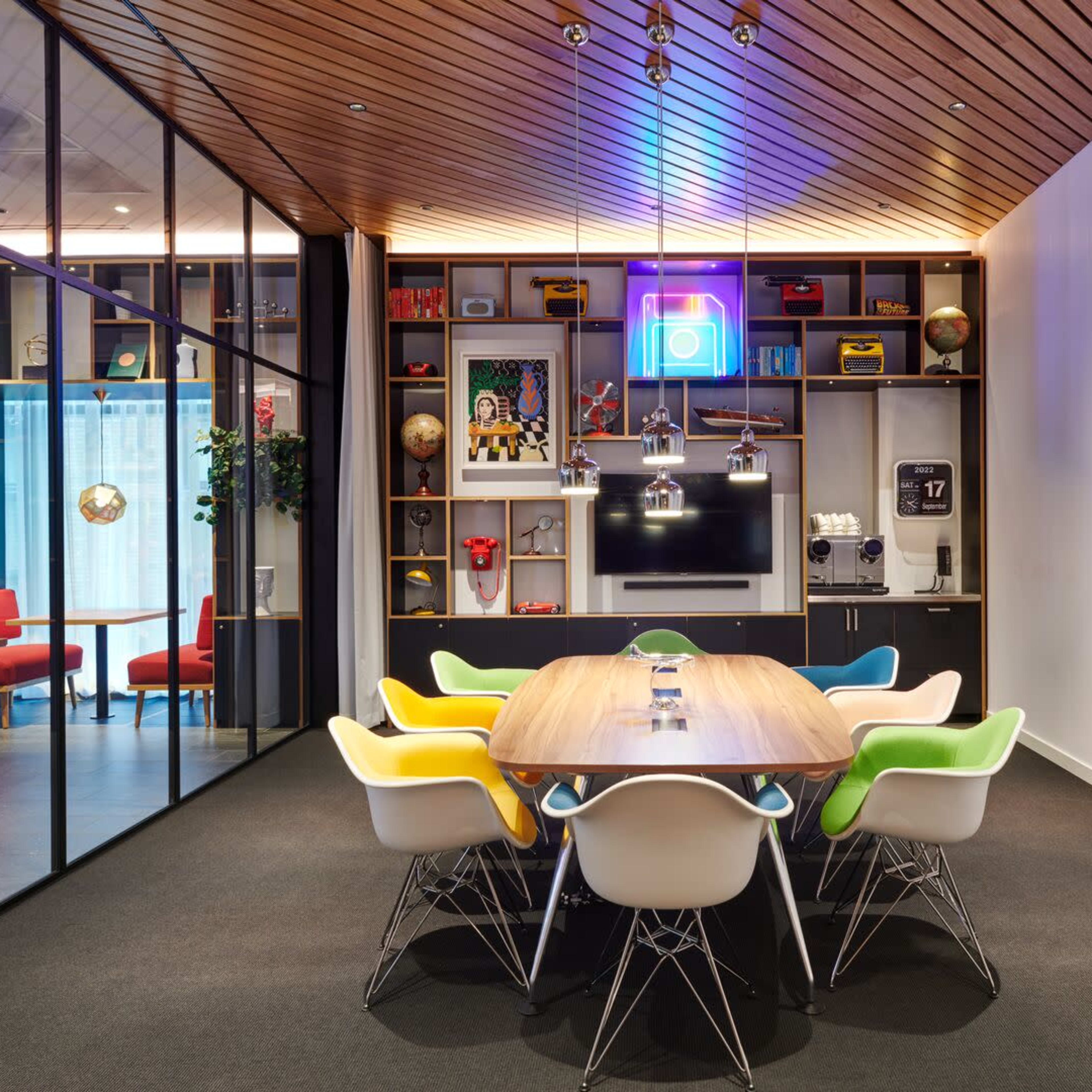 A modern conference room features a large wooden table surrounded by colorful chairs, with shelves displaying various decor items and a television mounted on the wall.