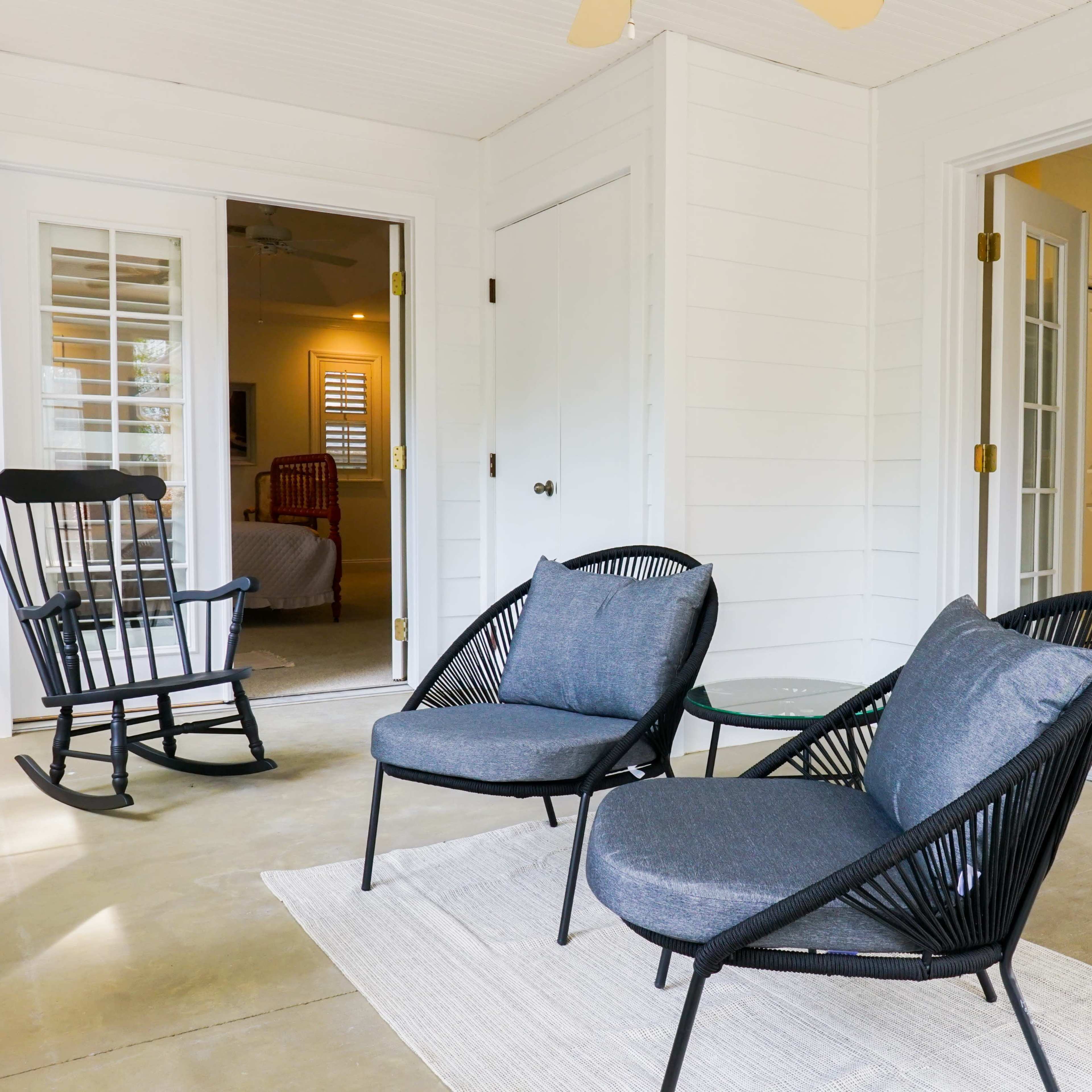 A covered porch with two black lounge chairs, a small glass table, and a rocking chair, all arranged near an entrance to a kitchen.