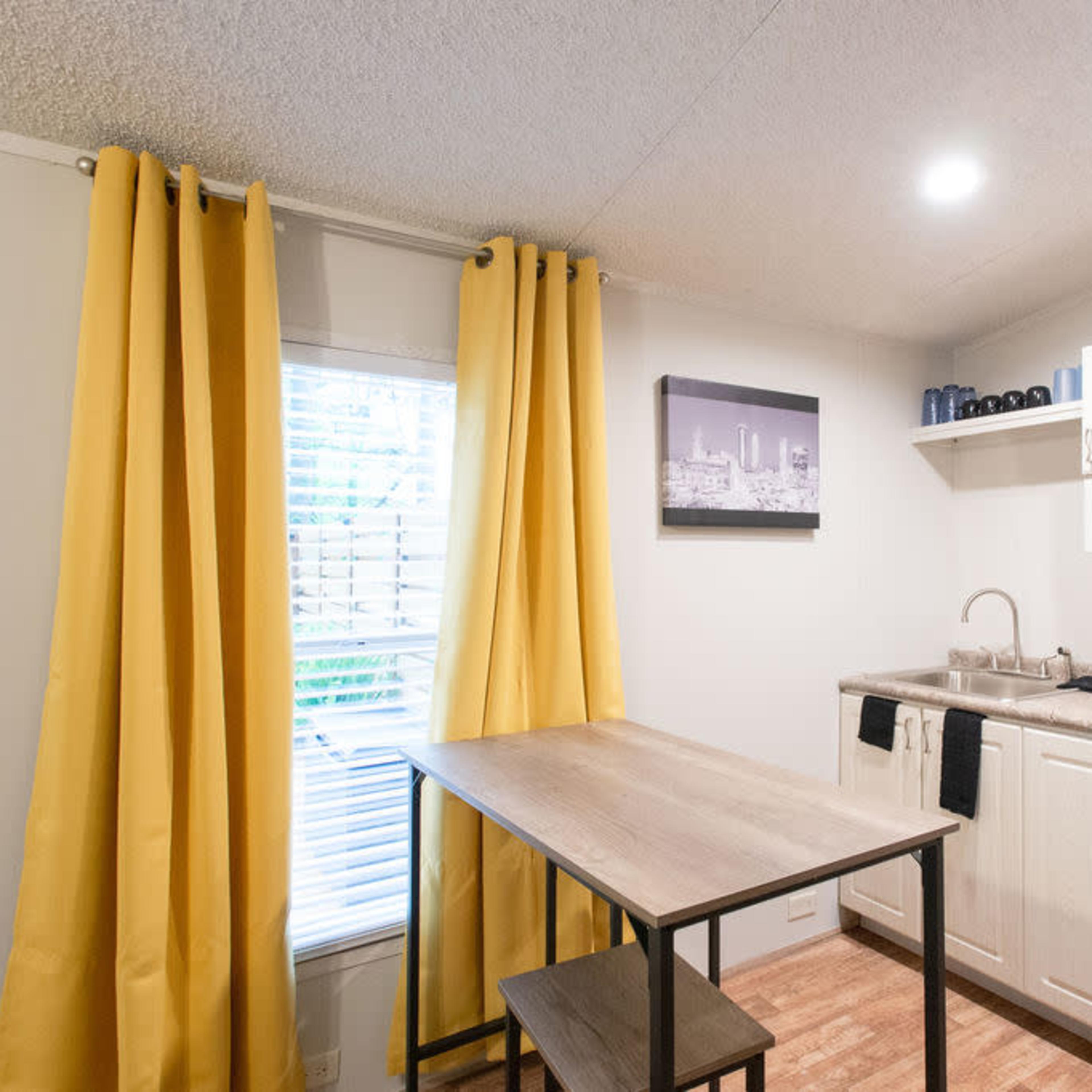 A small kitchen area with a table, two chairs, yellow curtains, and a wall-mounted air conditioning unit.
