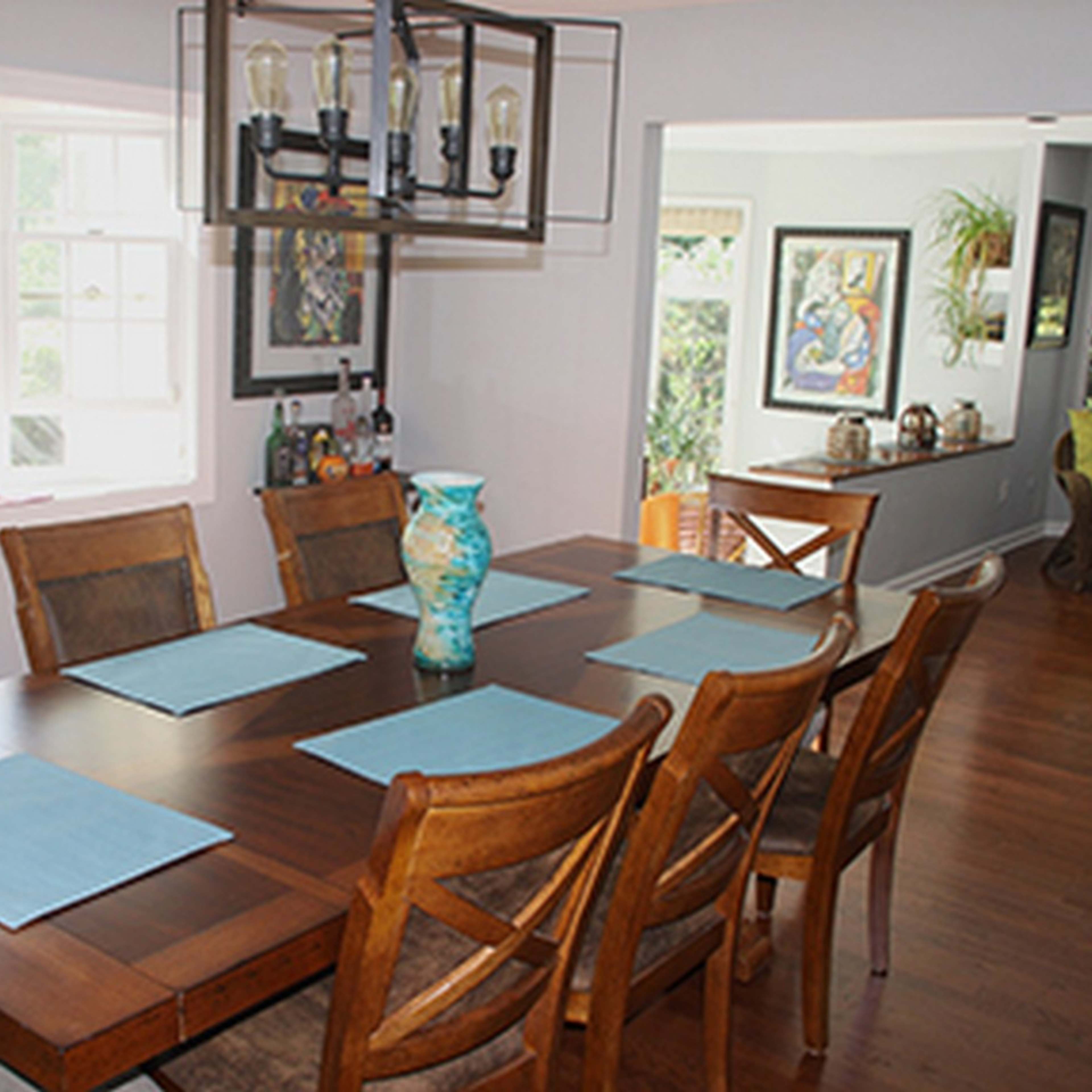 A spacious dining room features a large wooden table set with blue placemats and a decorative vase, surrounded by wooden chairs and illuminated by a modern chandelier.