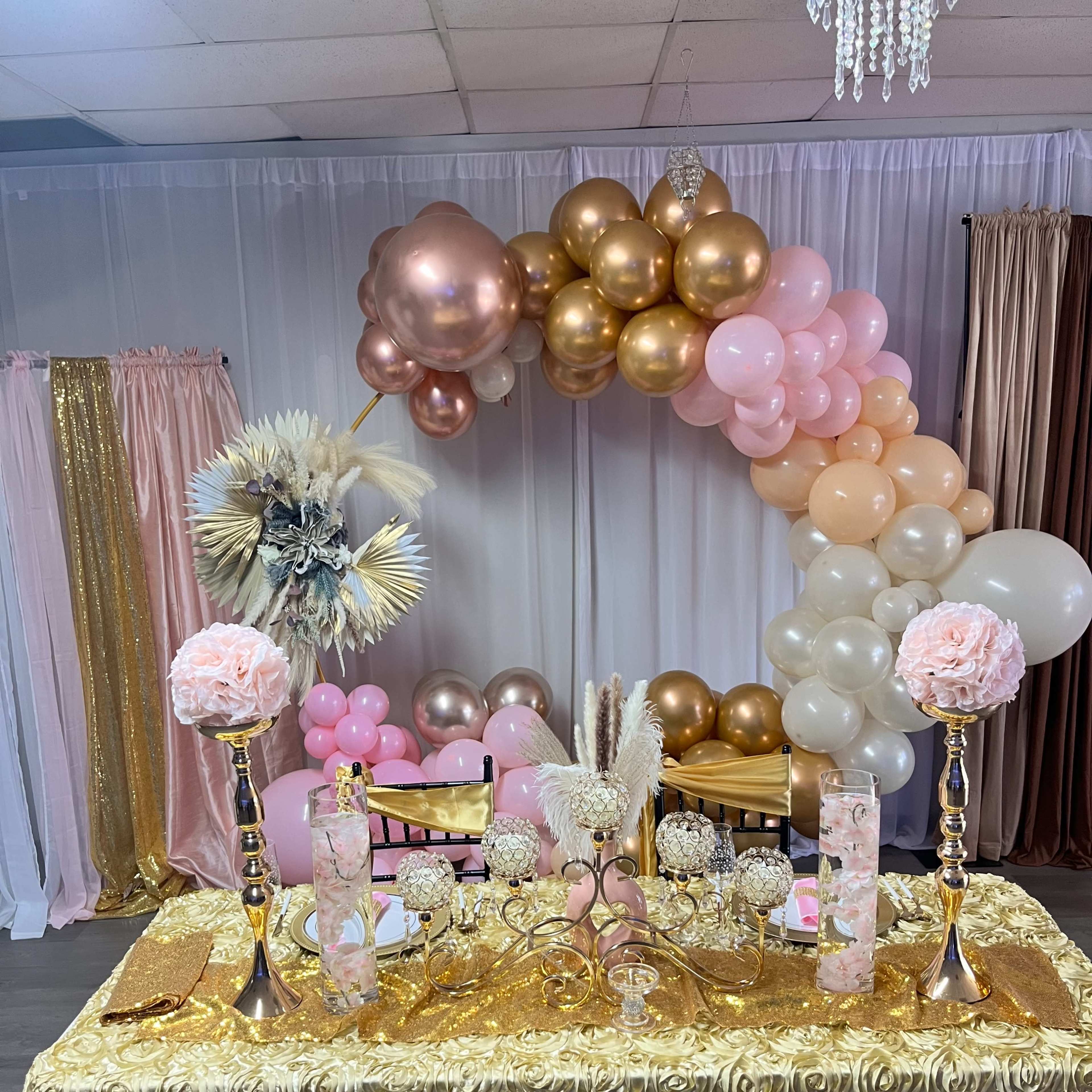 A decorated table with a gold tablecloth adorned with floral arrangements, elegant candles, and a colorful balloon arch in the background.