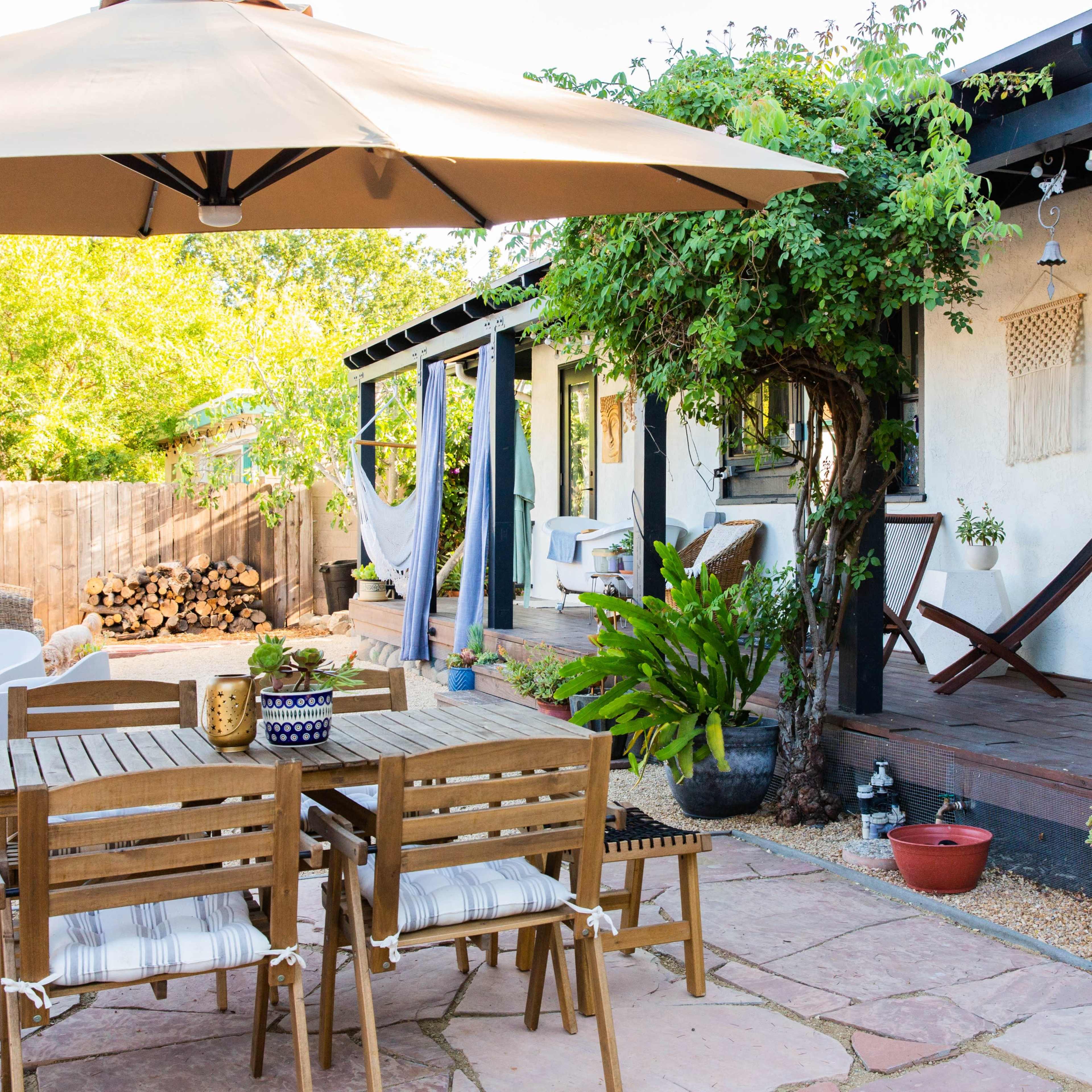 A patio area features a wooden dining table with chairs, shaded by an umbrella, surrounded by potted plants and a firewood stack in the background.