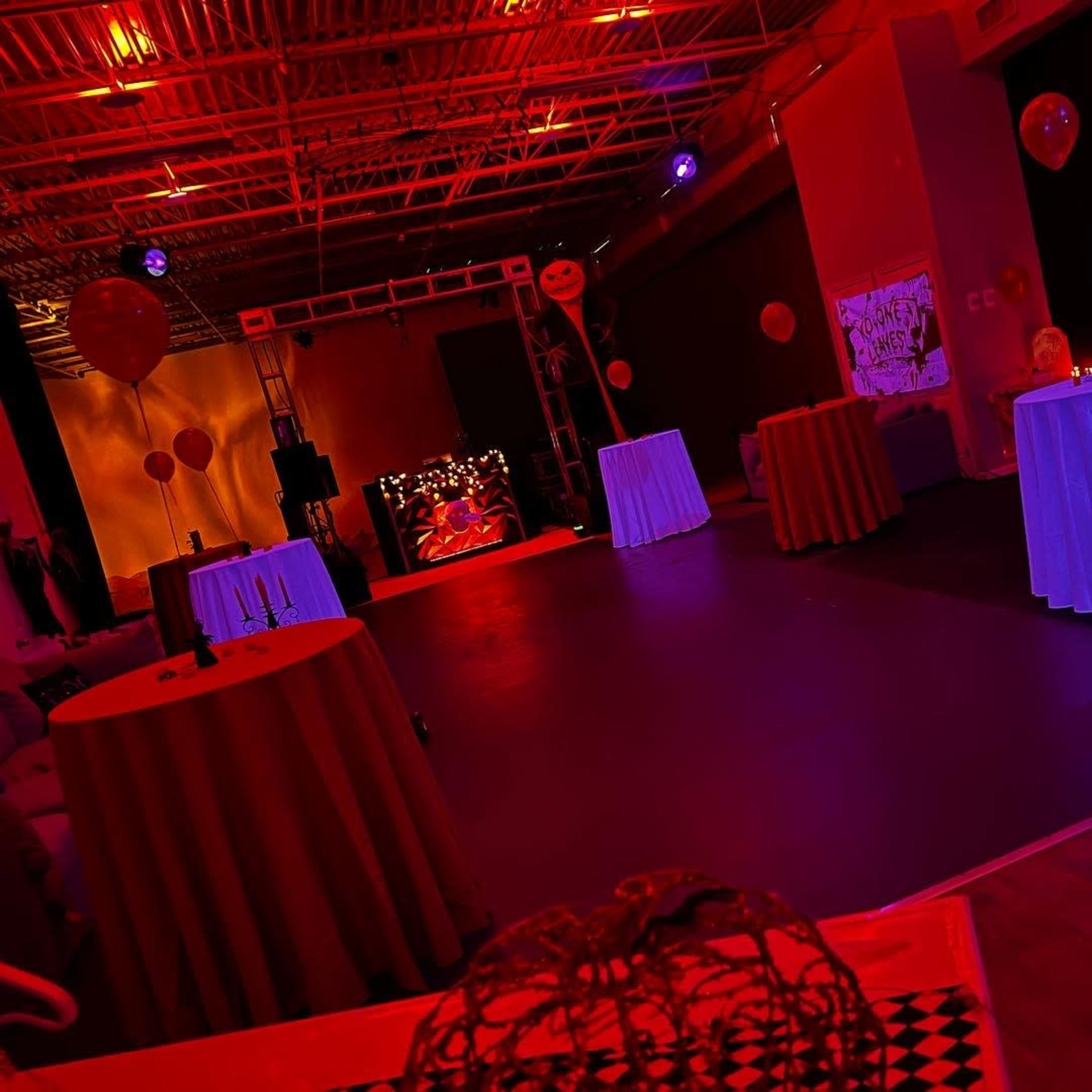 A dimly lit event space decorated with round tables and red lighting, featuring balloon decorations and a stage at the far end.