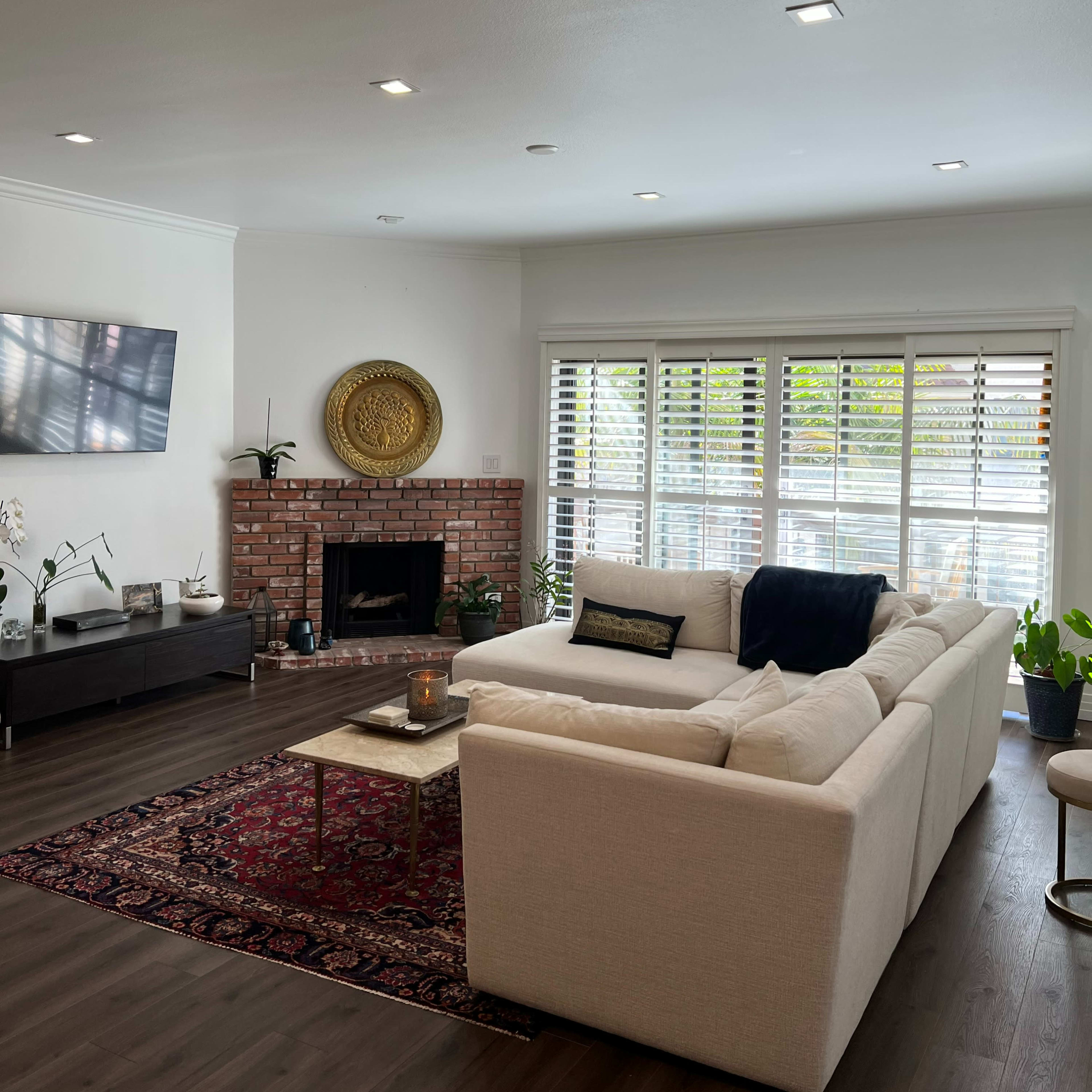 A modern living room with a sectional sofa, a brick fireplace, and large windows covered with wooden shutters.