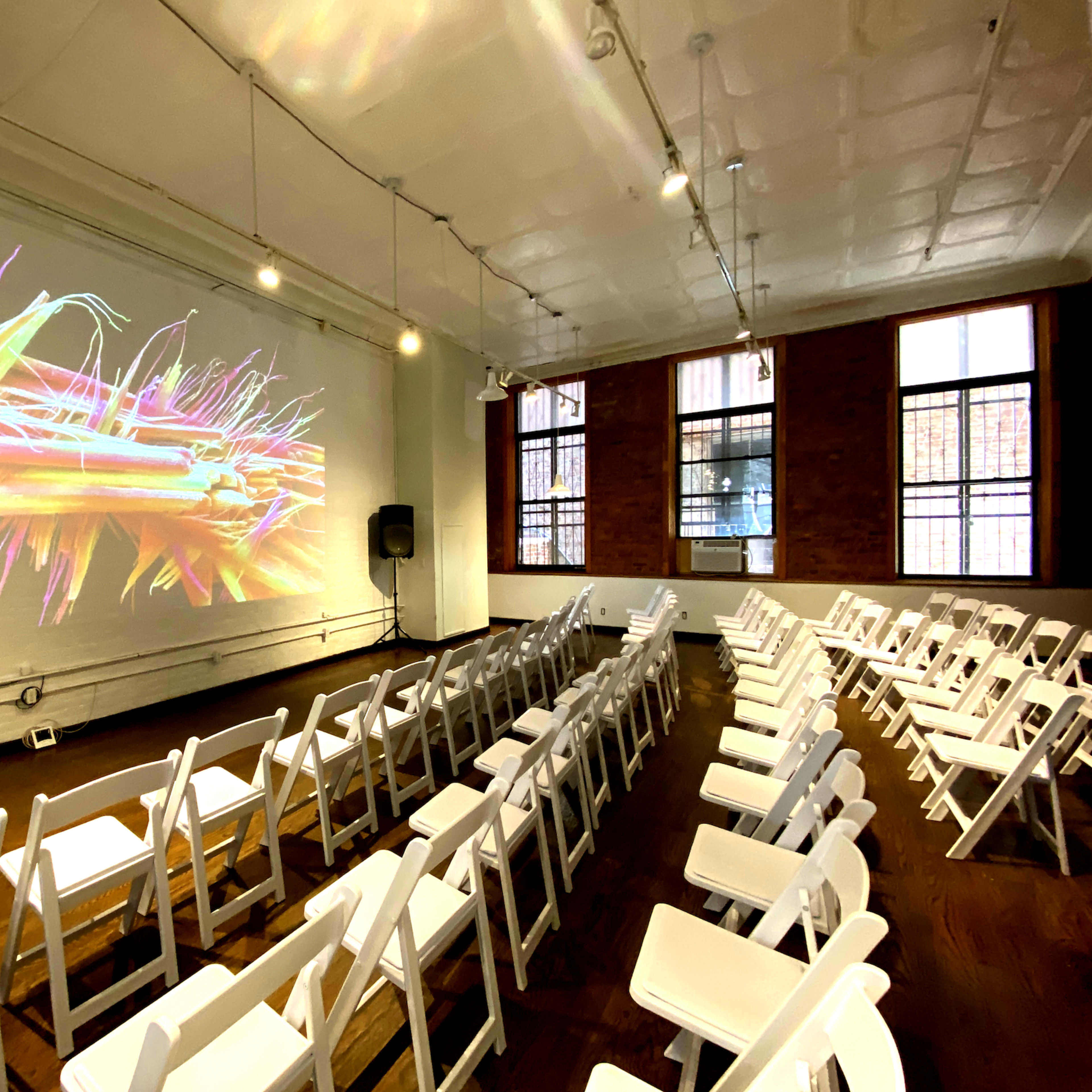A spacious room with rows of white folding chairs facing a colorful projection on a wall, flanked by large windows.