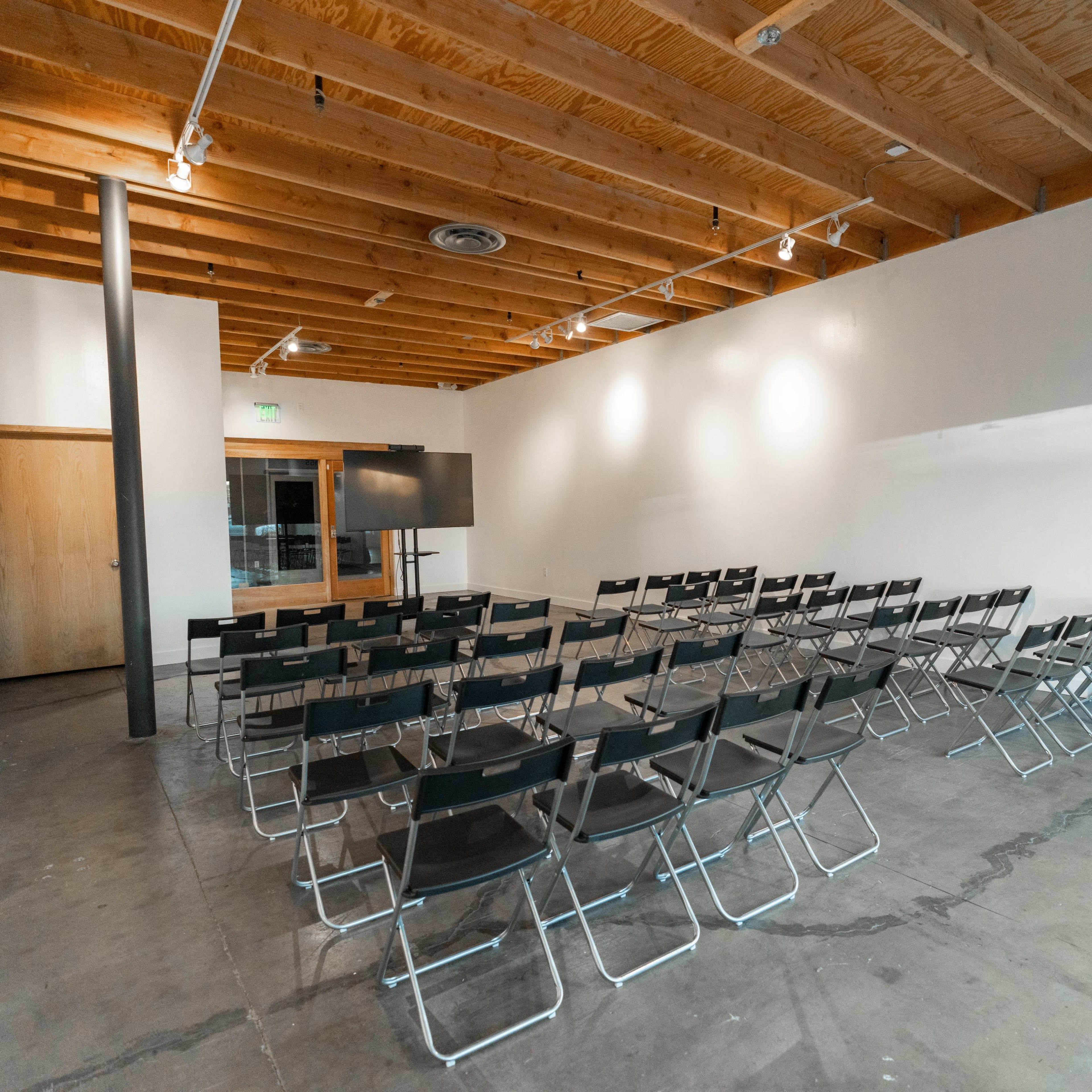 A row of black folding chairs is arranged facing a blank wall in a sparsely furnished meeting room with wooden beams.