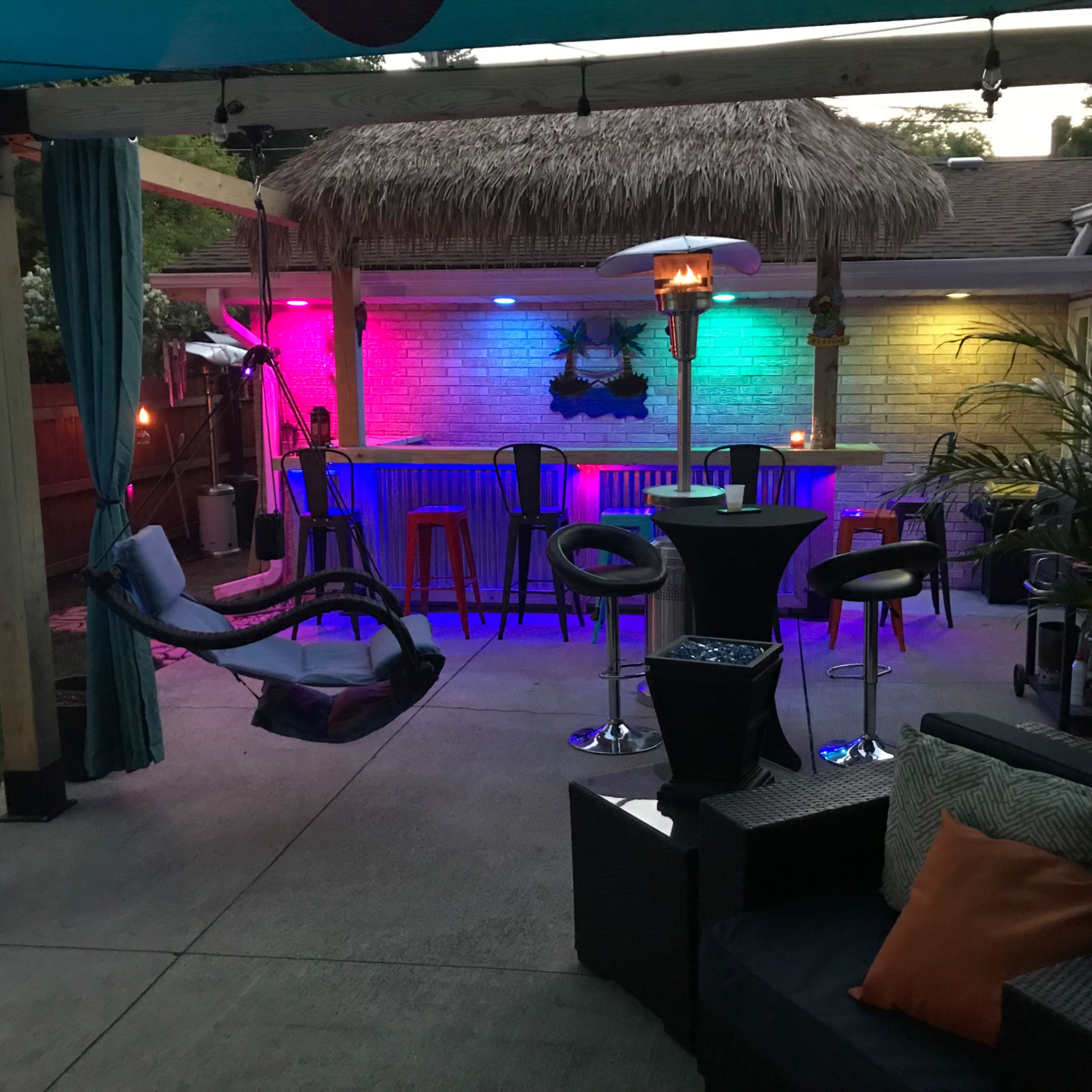 A backyard patio features a thatched-roof bar with colorful LED lighting, surrounded by seating areas and a hammock.