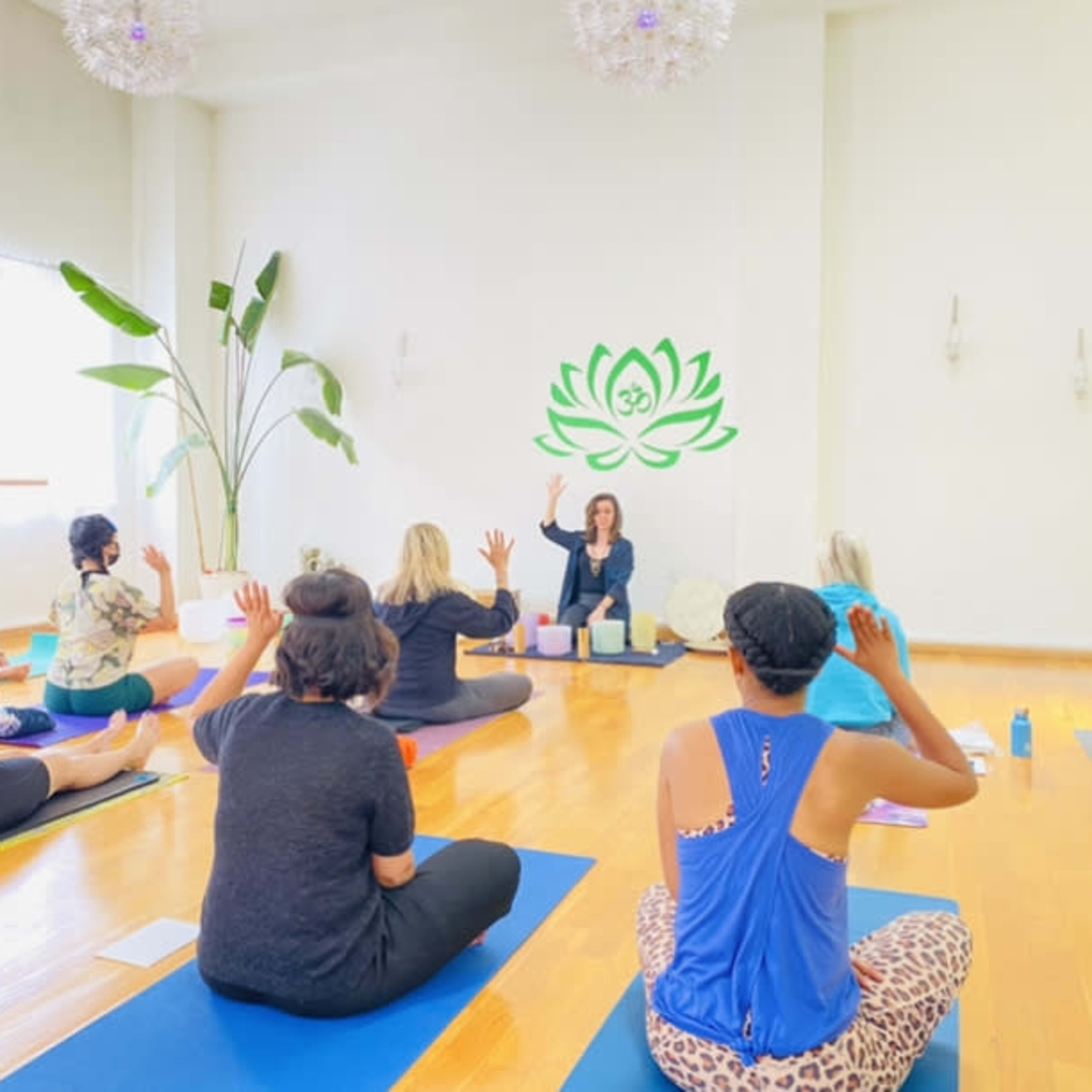A group of people sits on yoga mats in a bright studio, attentively facing an instructor who is leading a class.