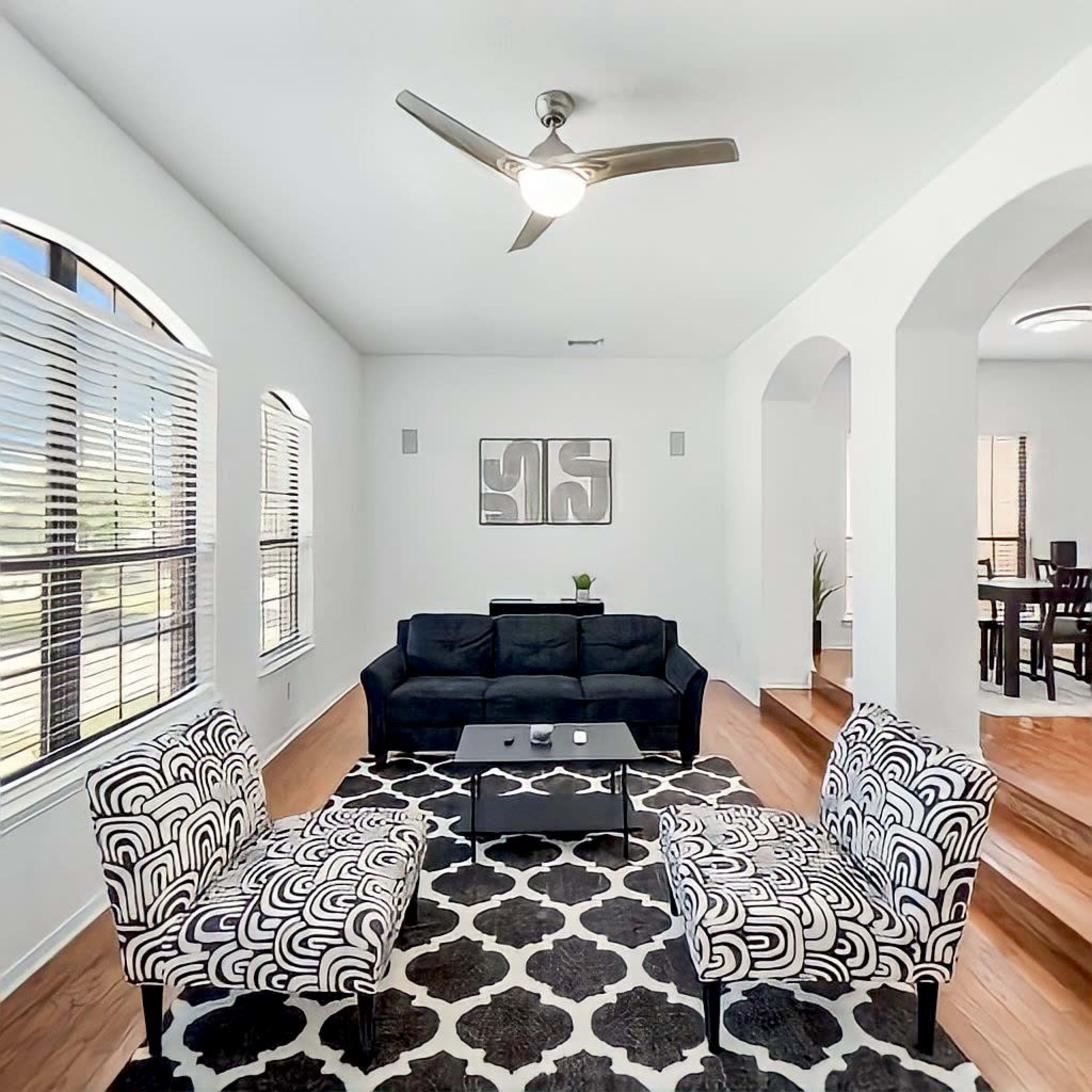 A modern living room features a black sofa, two patterned armchairs, a coffee table, and large windows providing natural light.