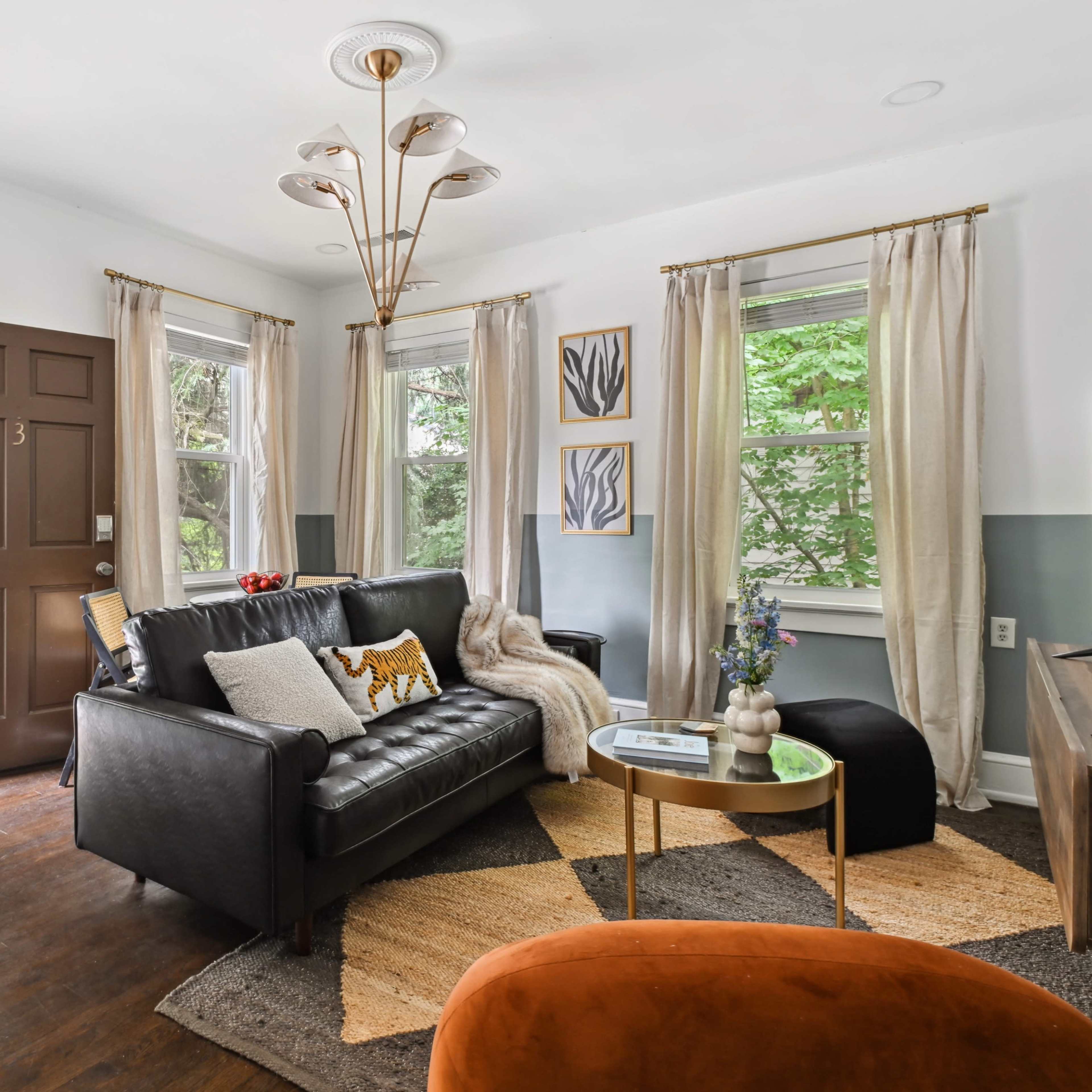 The living room features a black leather sofa, a round coffee table, and large windows that overlook a tree-filled balcony.