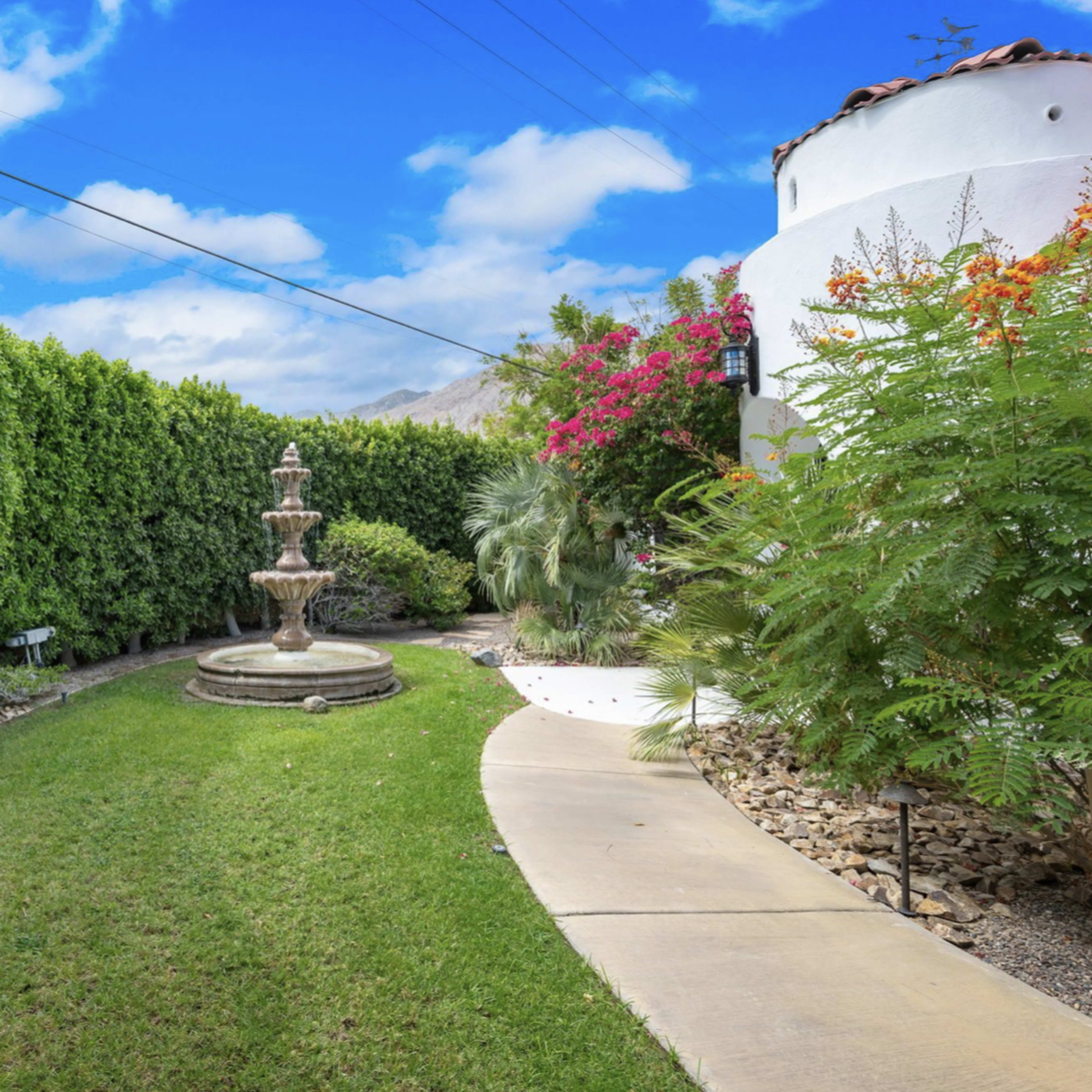 A landscaped garden featuring a fountain surrounded by trimmed hedges and colorful plants under a blue sky.