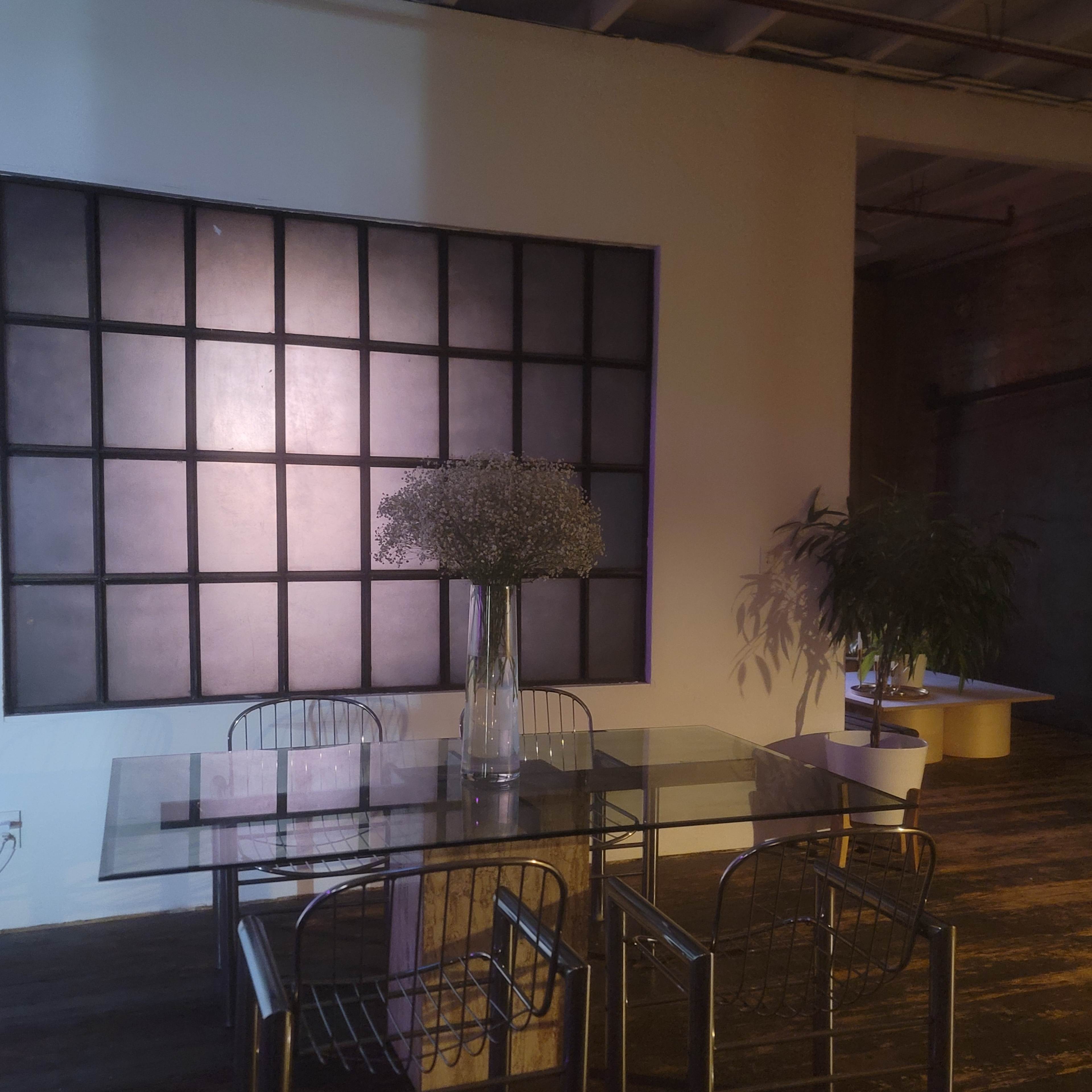 A glass table with metal chairs is situated in a dimly lit room featuring a large grid-patterned wall and a tall vase of dried flowers.