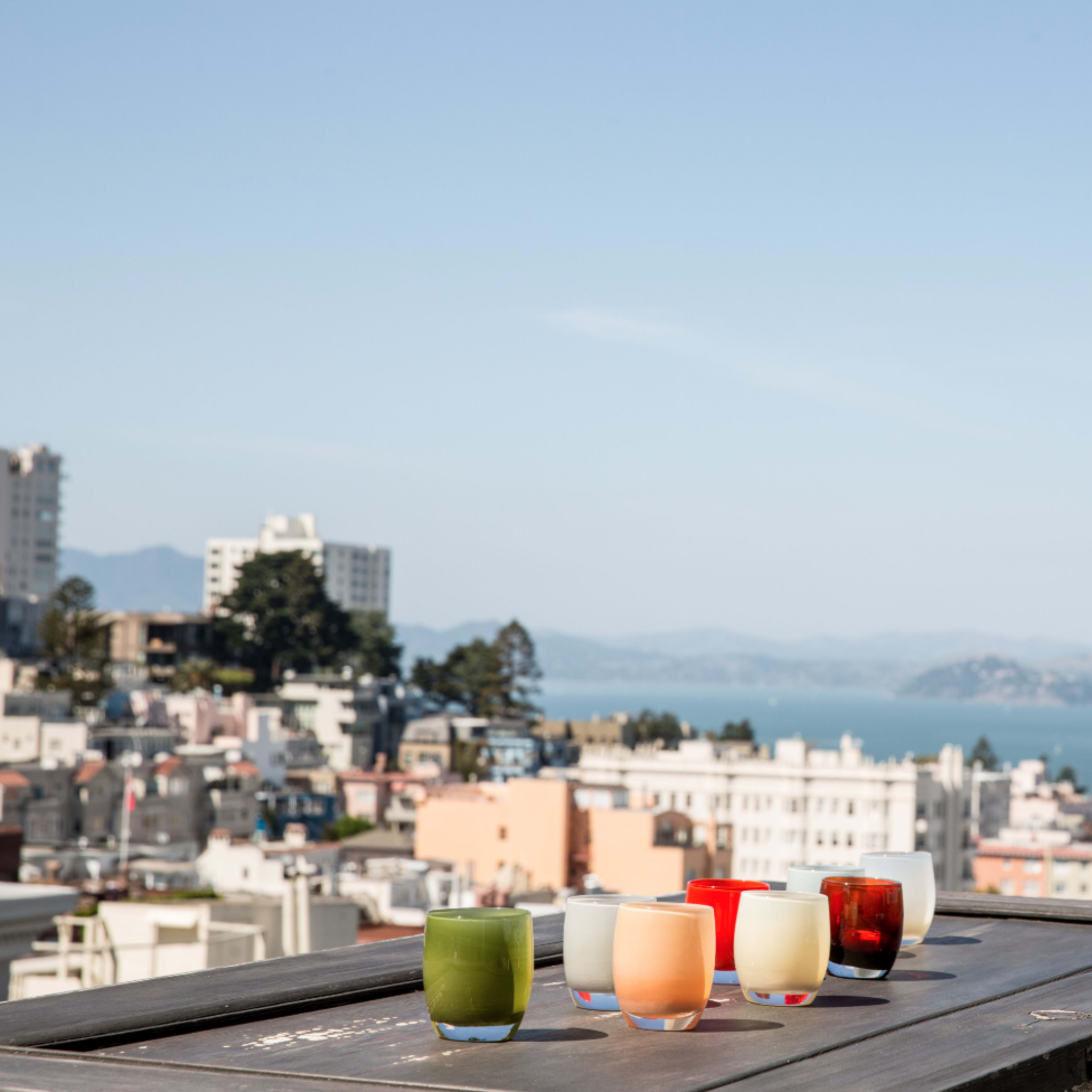 Six colorful glasses are arranged on a wooden table, overlooking a coastal city and body of water in the background.