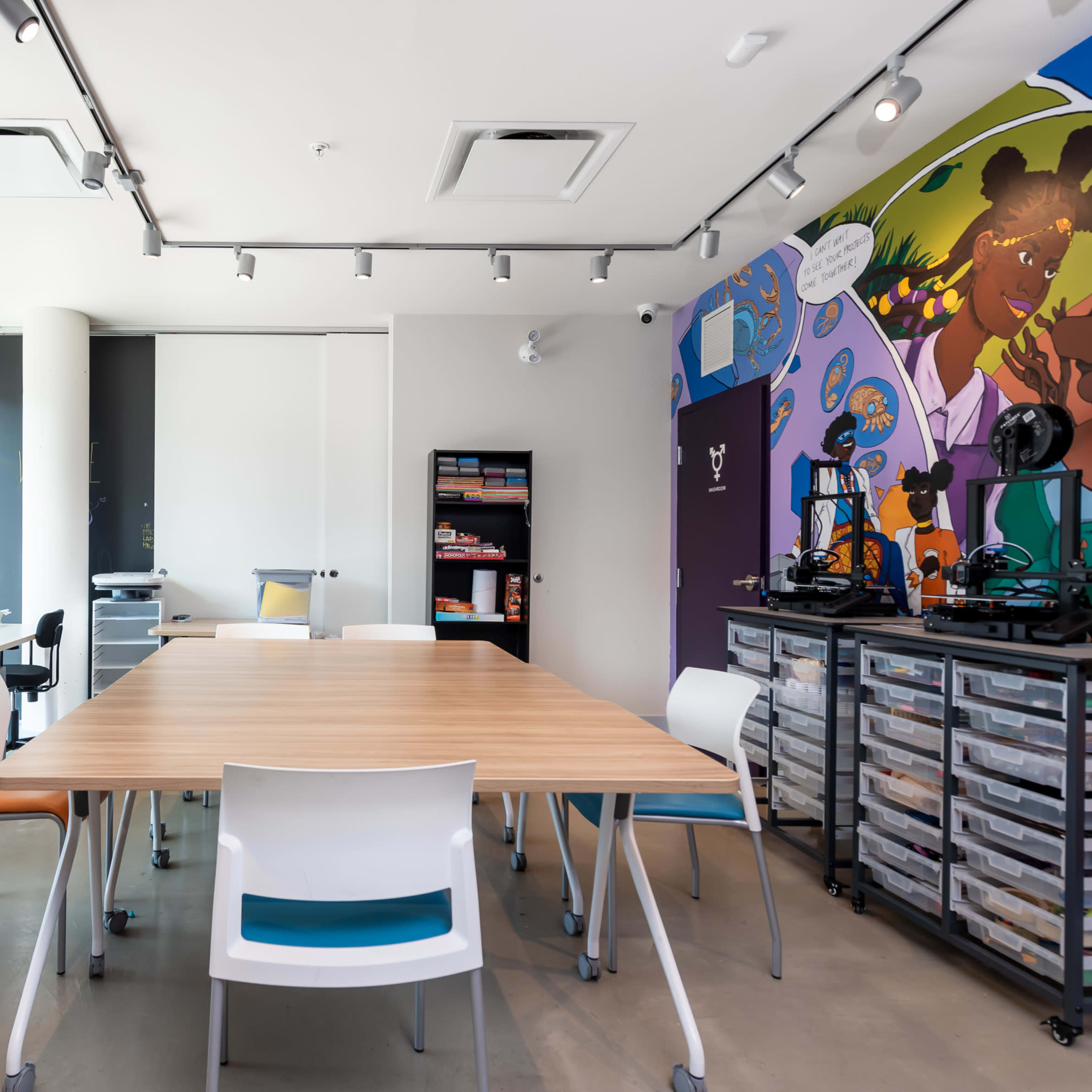 A spacious, modern office features a large wooden table surrounded by colorful chairs, with large windows letting in natural light and a mural on the wall.