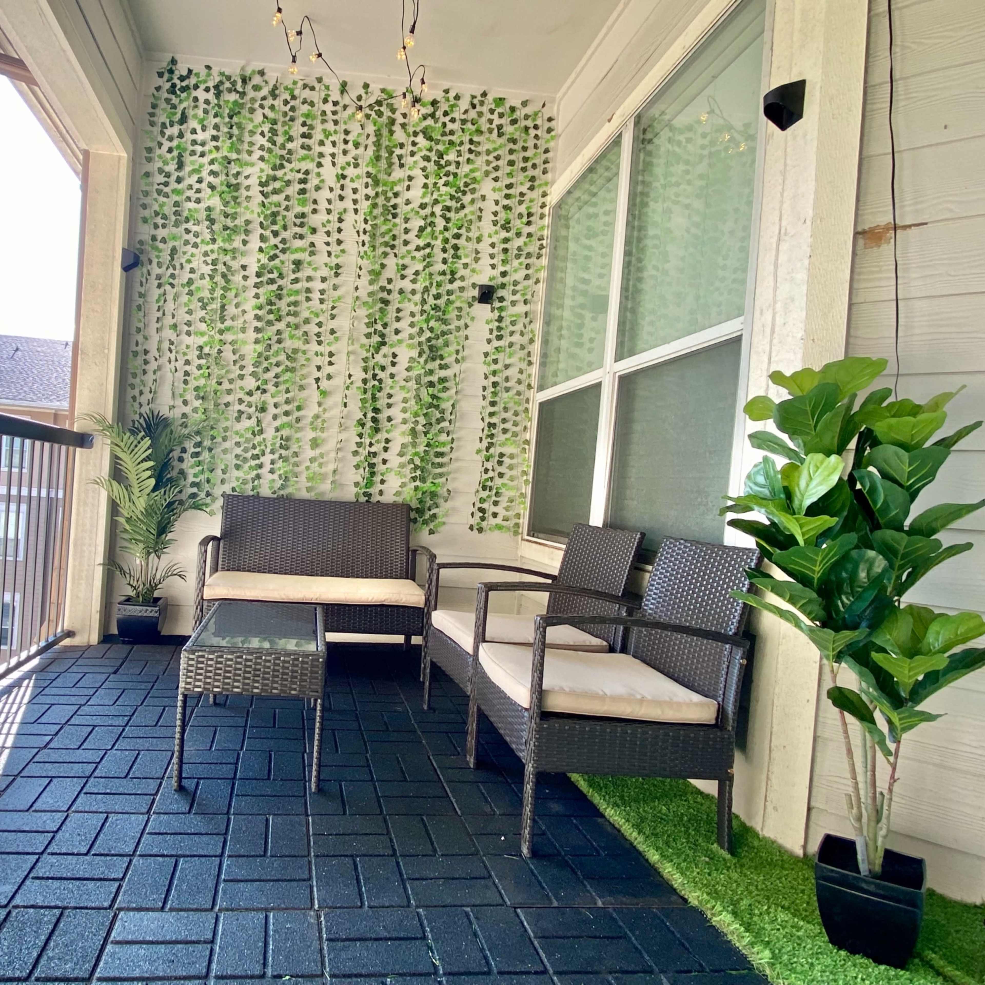 A patio area features wicker furniture with cushions, a green wall decoration, and potted plants, set against a backdrop of an apartment complex.