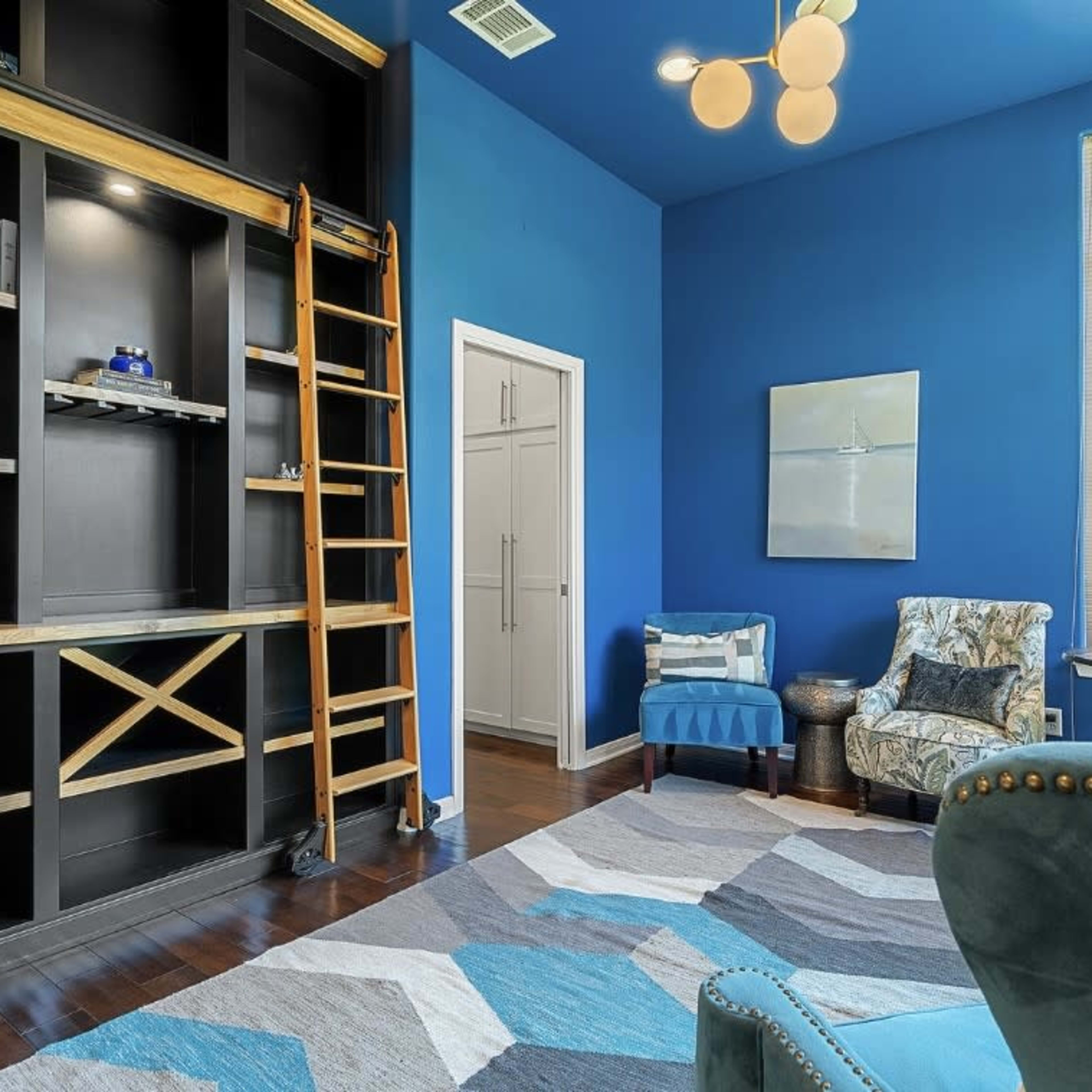 A modern room with blue walls, a wooden ladder leading to a tall bookshelf, and a patterned rug on the floor.