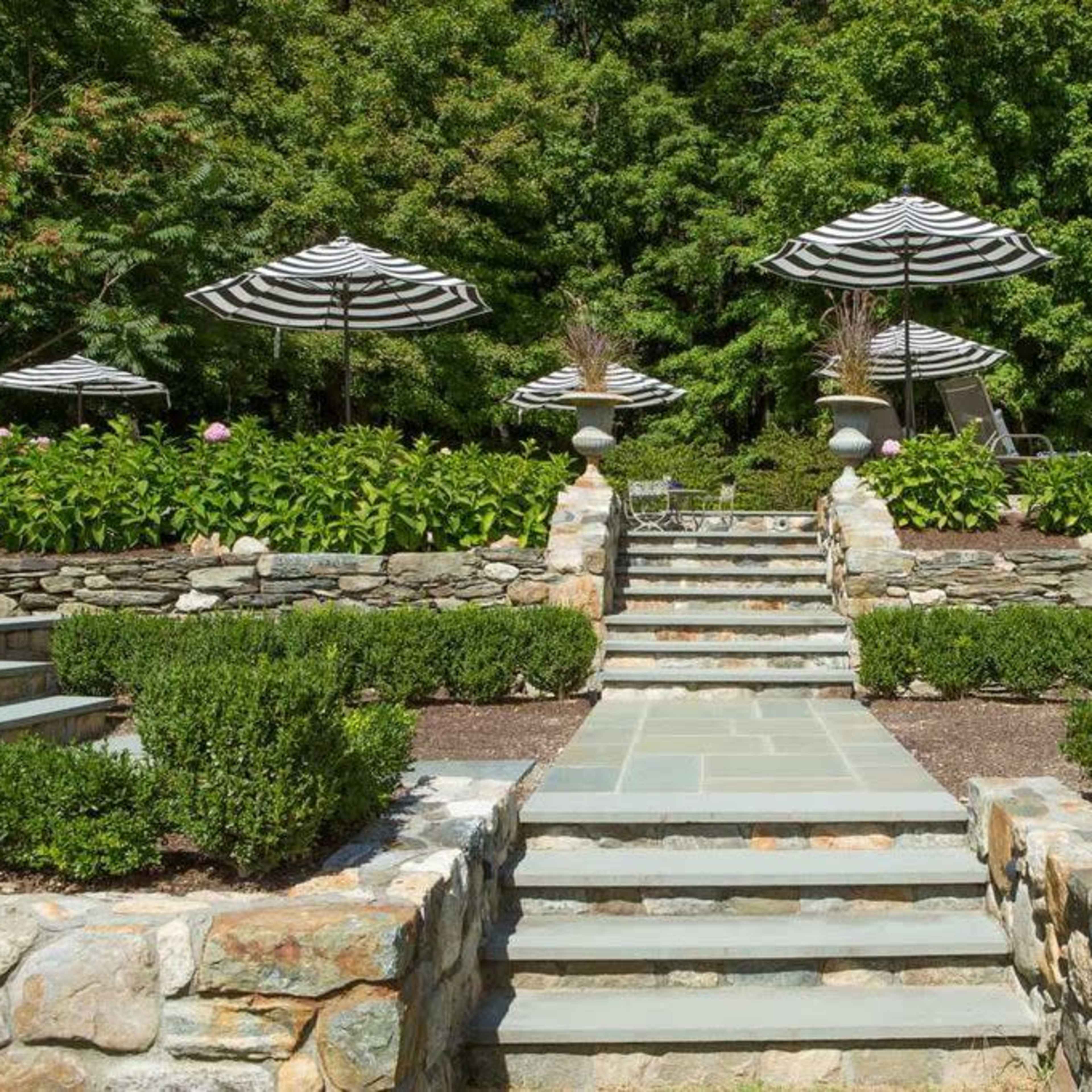 A landscaped garden with stone steps leading up to a terrace featuring striped umbrellas and neatly trimmed hedges.
