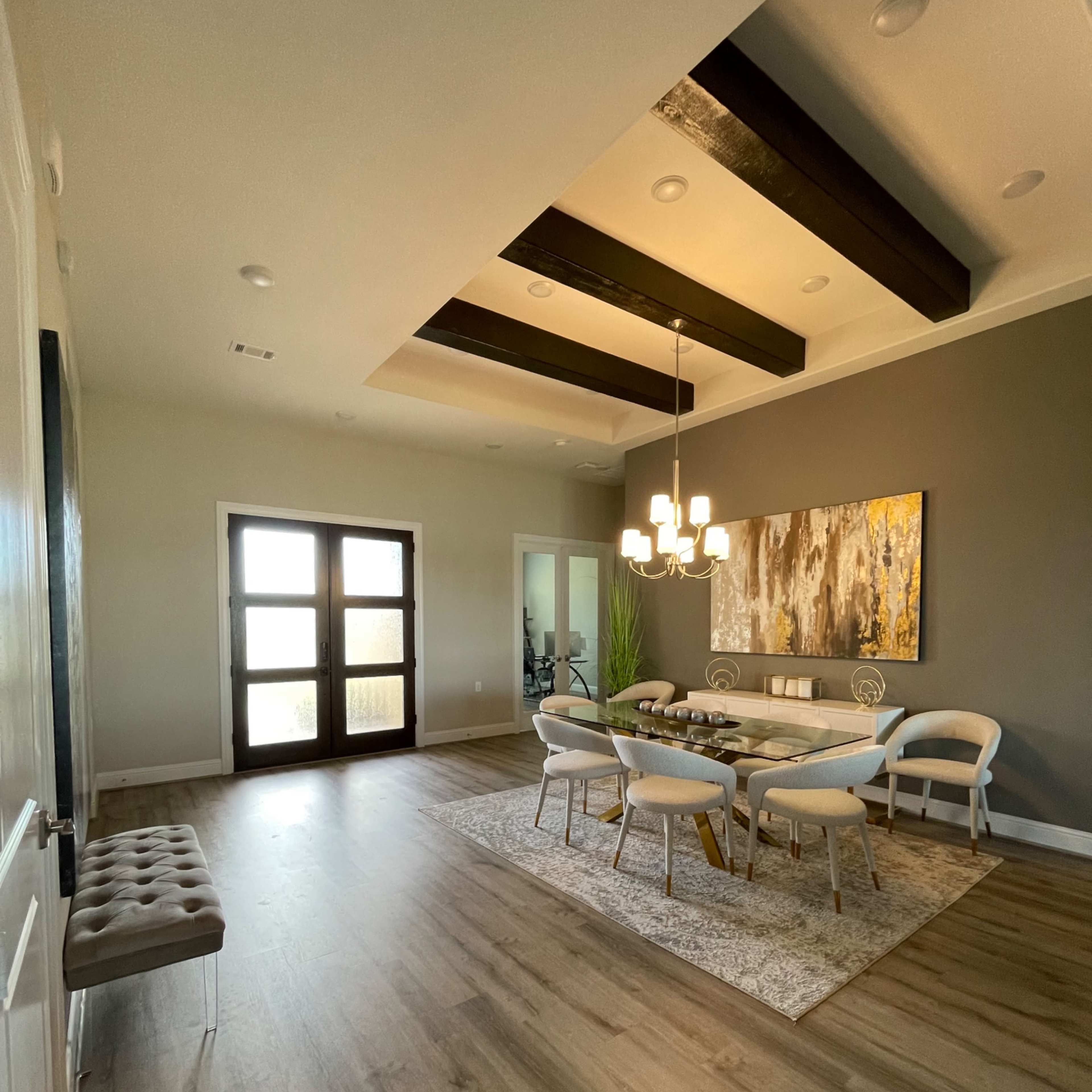 A dining area features a round table surrounded by chairs, with large windows and a coffered ceiling illuminated by a modern chandelier.
