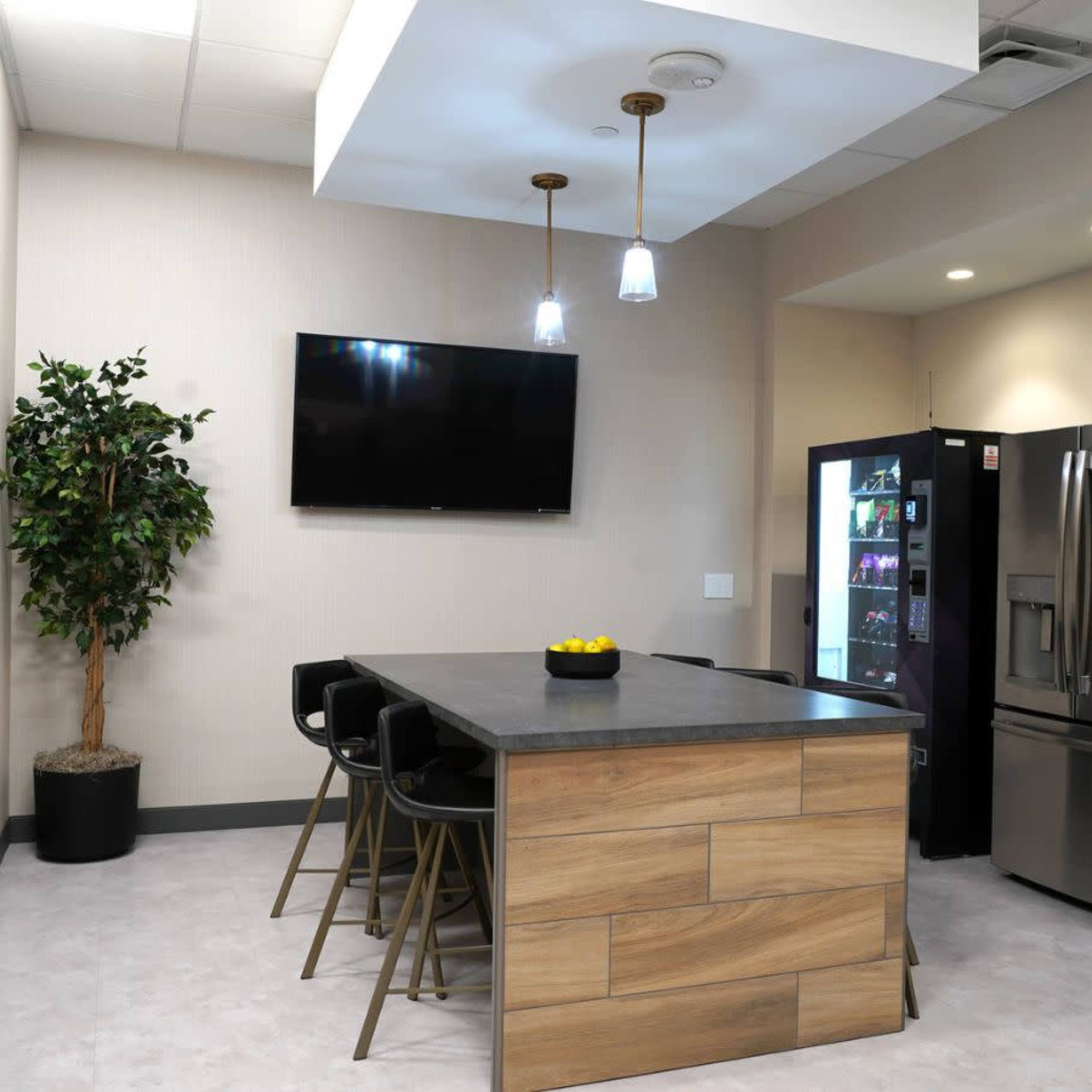 A modern kitchen area with a central island, four black bar stools, a refrigerator, a television mounted on the wall, and a small indoor plant.