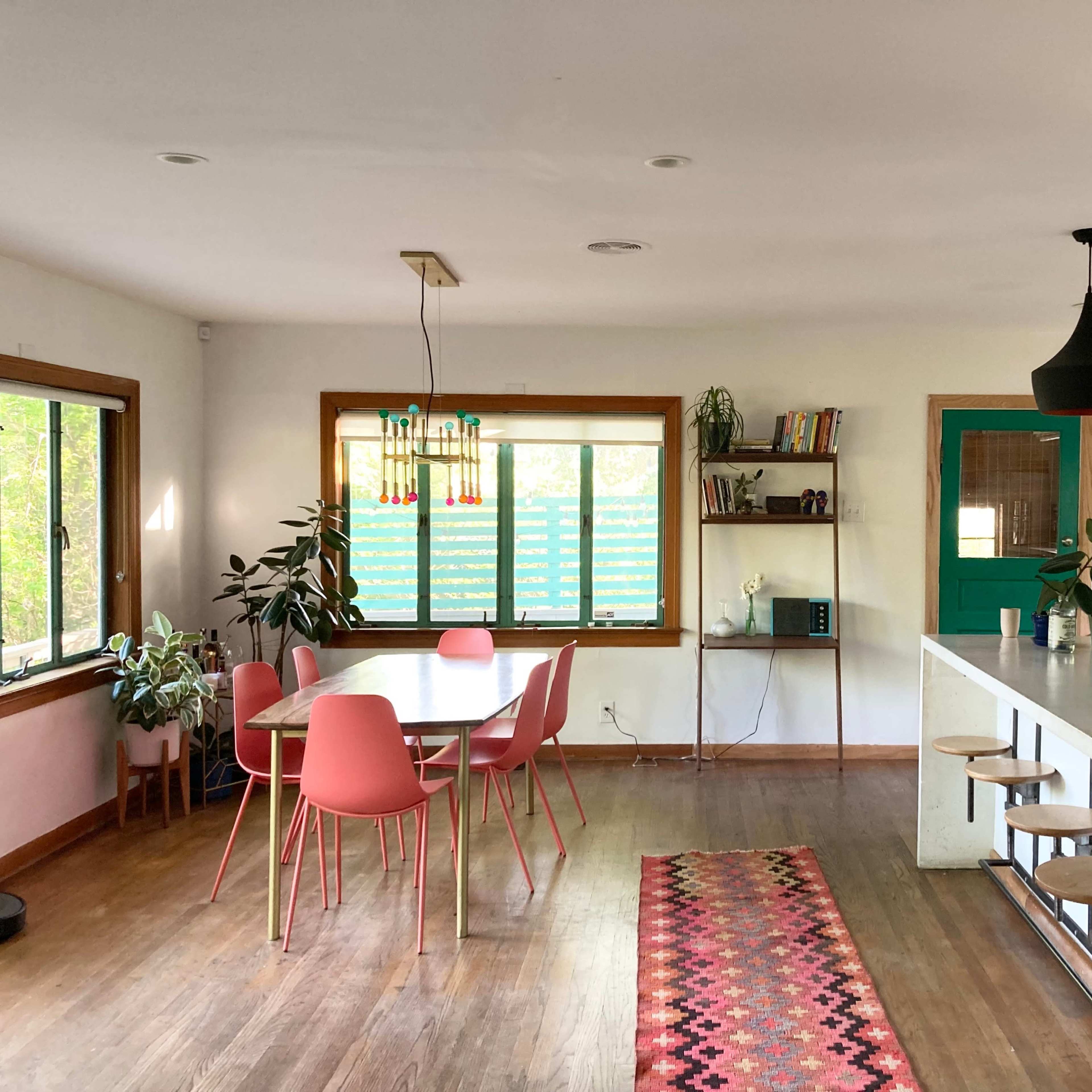 A modern kitchen and dining area featuring a round table with pink chairs, a long bar with stools, and large windows allowing natural light.