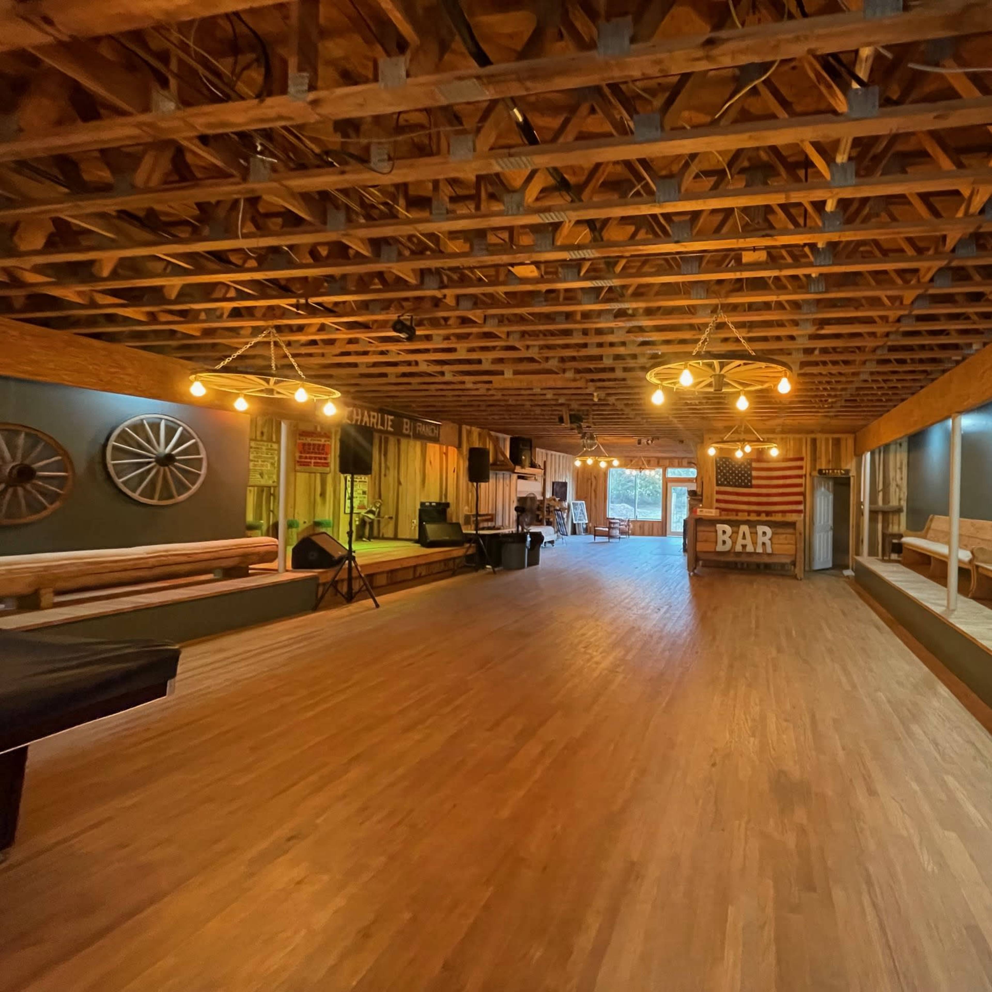A spacious interior with wooden flooring, exposed beams, and a bar area in the corner, illuminated by hanging light fixtures.