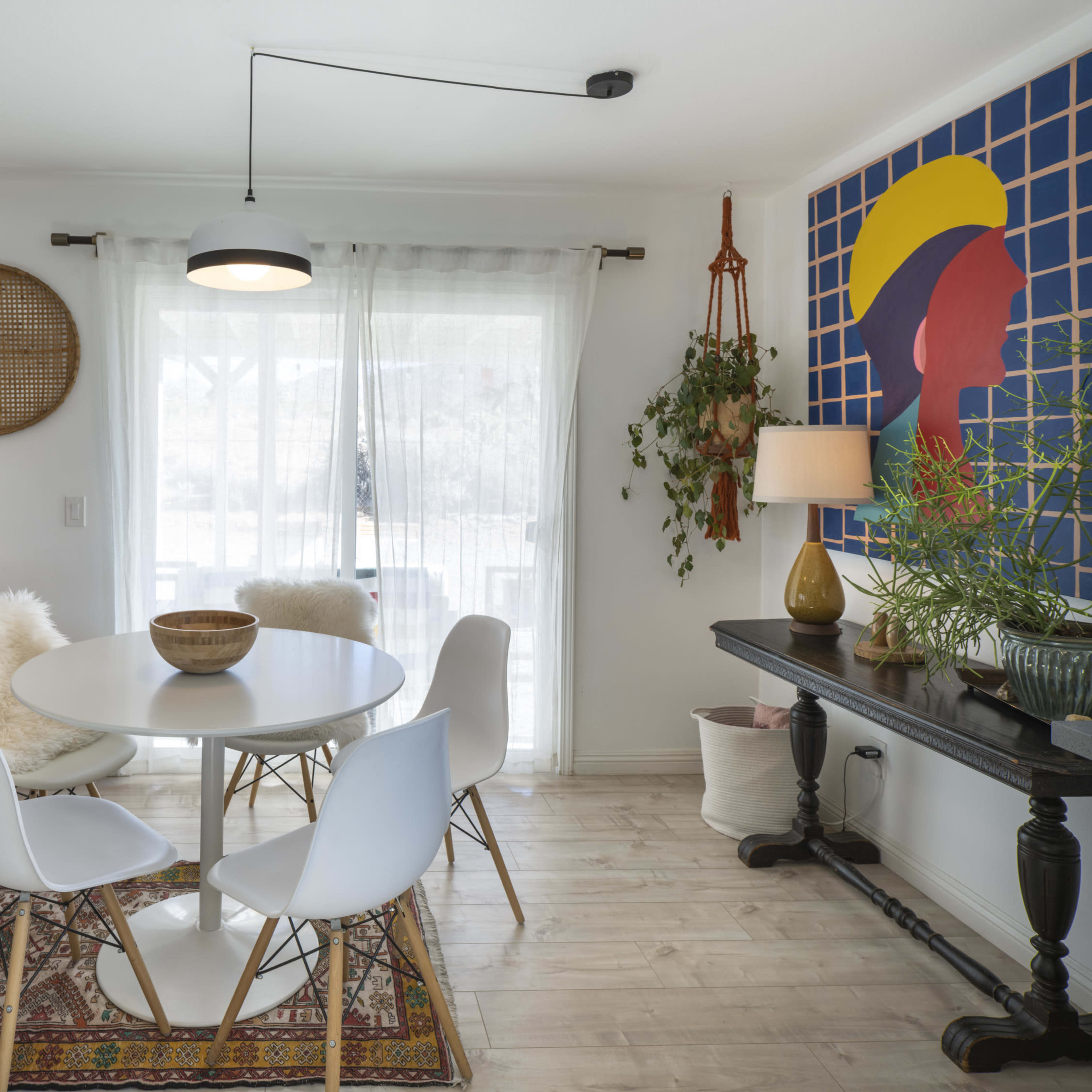A modern dining area features a round table with white chairs, a patterned rug, and a large wall art piece against a bright window backdrop.